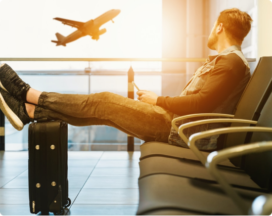 A man sitting in an airport resting his feet on a suitcase while watching a plane take off in the distance