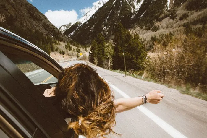 The view of the back of a woman with long brown hair hanging her head and arm out of a car window looking at mountains in the distance