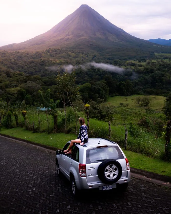 A young woman sits on the roof of a silver SUV in front of a lush green field in front of a volcano in the distance