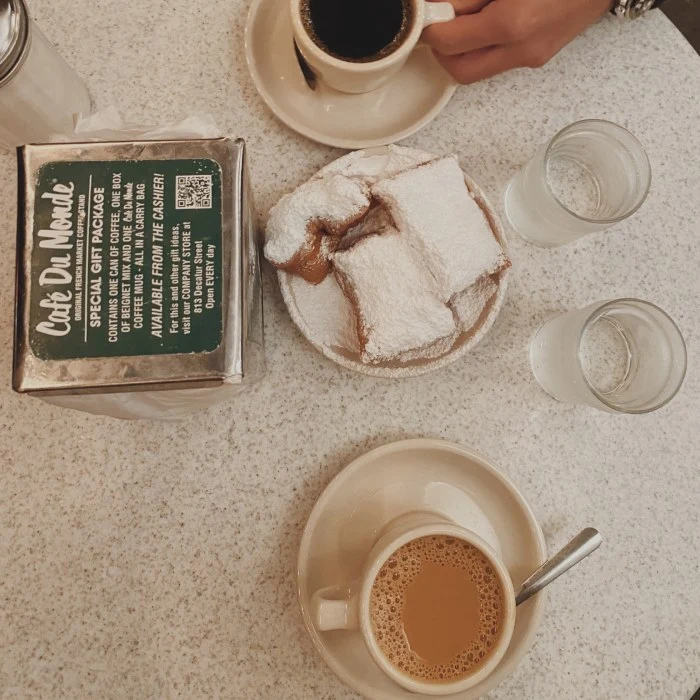 Two cups of coffee and a plate with three powdered beignets sit on a table with a Cafe Du Monde sign