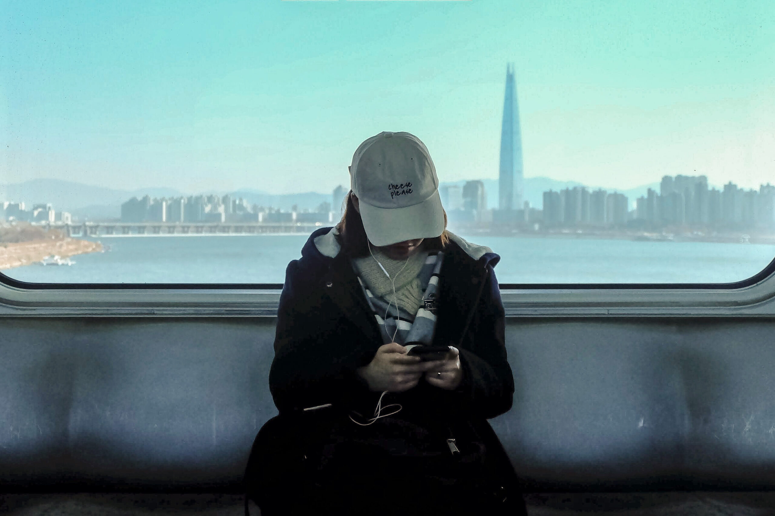 Student using smartphone on a train with the Seoul skyline in the background, showcasing everyday life in Korea.