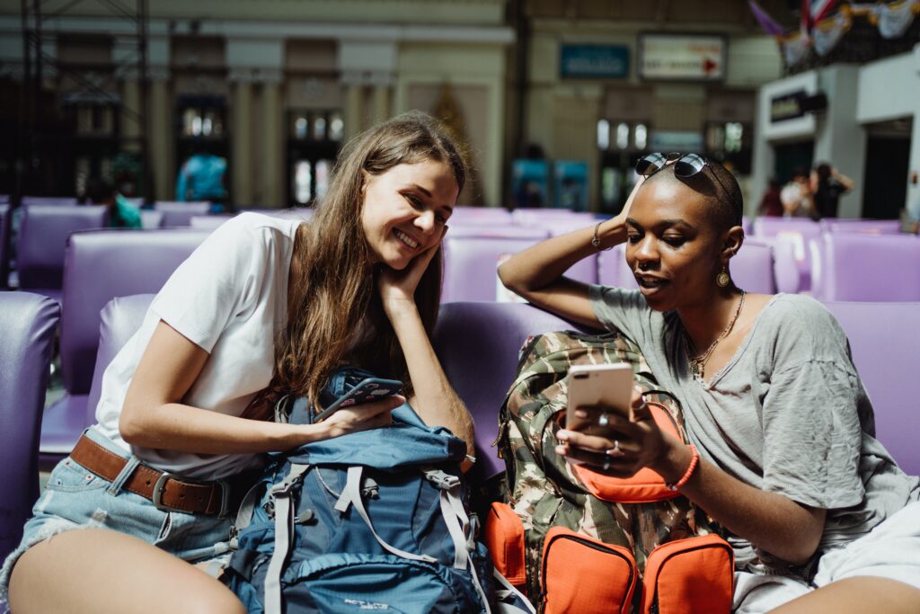 Two young women socializing in a train station while traveling. One woman is showing the other something on her phone and they are smiling.