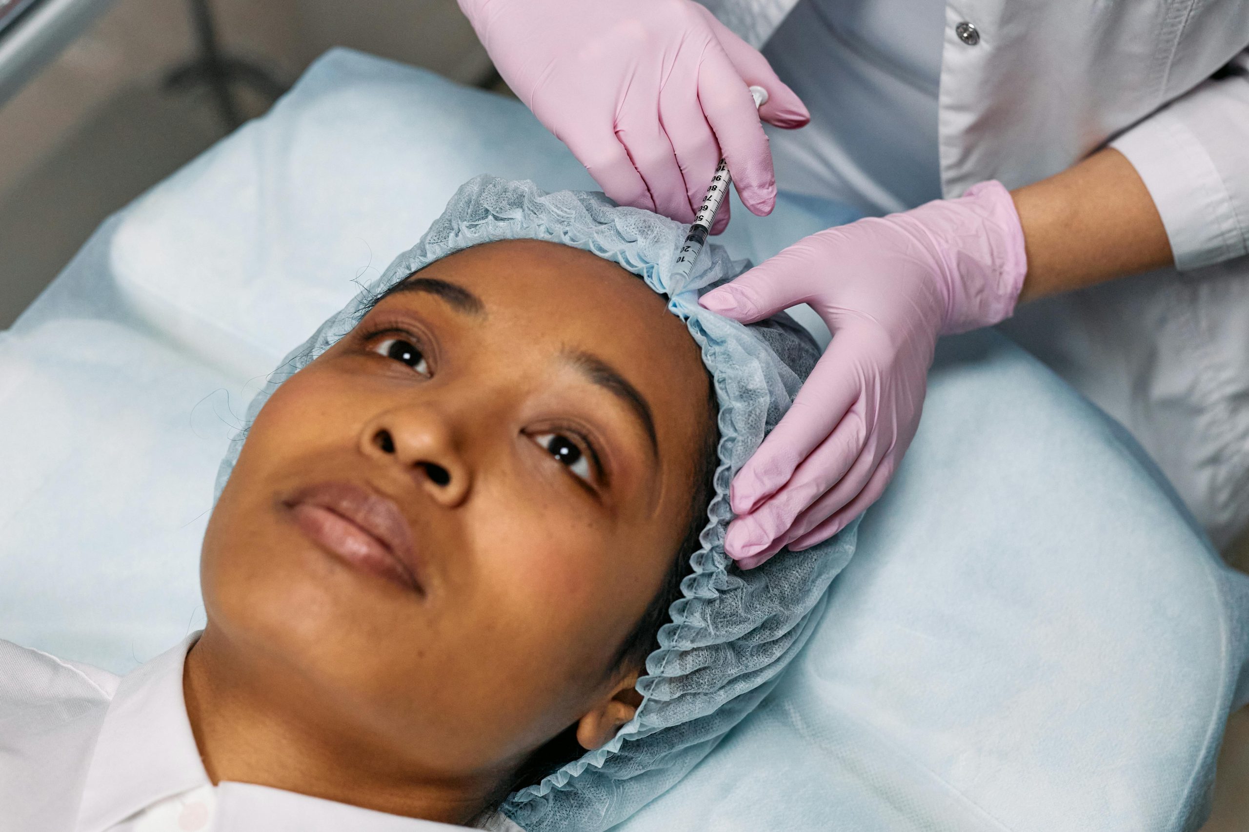 Close-up of a person receiving a cosmetic injection in the forehead while lying on a medical bed. The person is wearing a surgical cap, and a healthcare professional, wearing pink gloves, is administering the injection in a sterile environment.