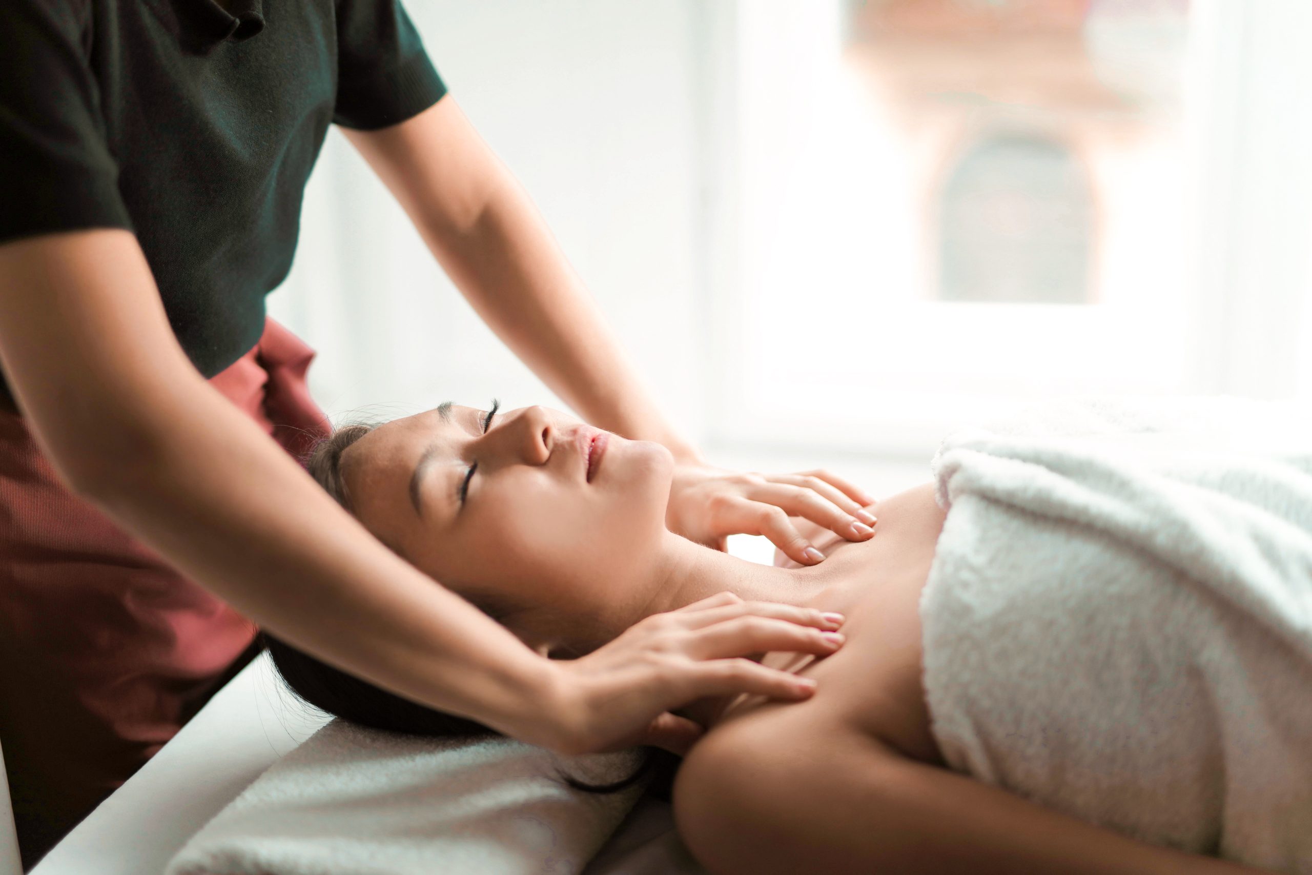 Person receiving a relaxing neck massage at a spa, lying down with eyes closed, covered in a white towel. The atmosphere is calm and peaceful, promoting wellness and relaxation.