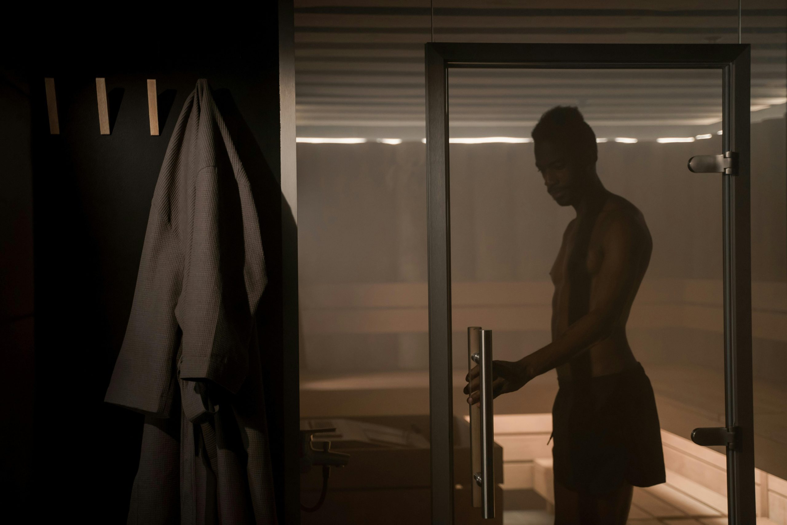 Person entering a dimly lit sauna, holding the door handle while wearing dark shorts. A robe hangs on a hook outside, and the warm, serene atmosphere suggests a moment of relaxation and self-care.