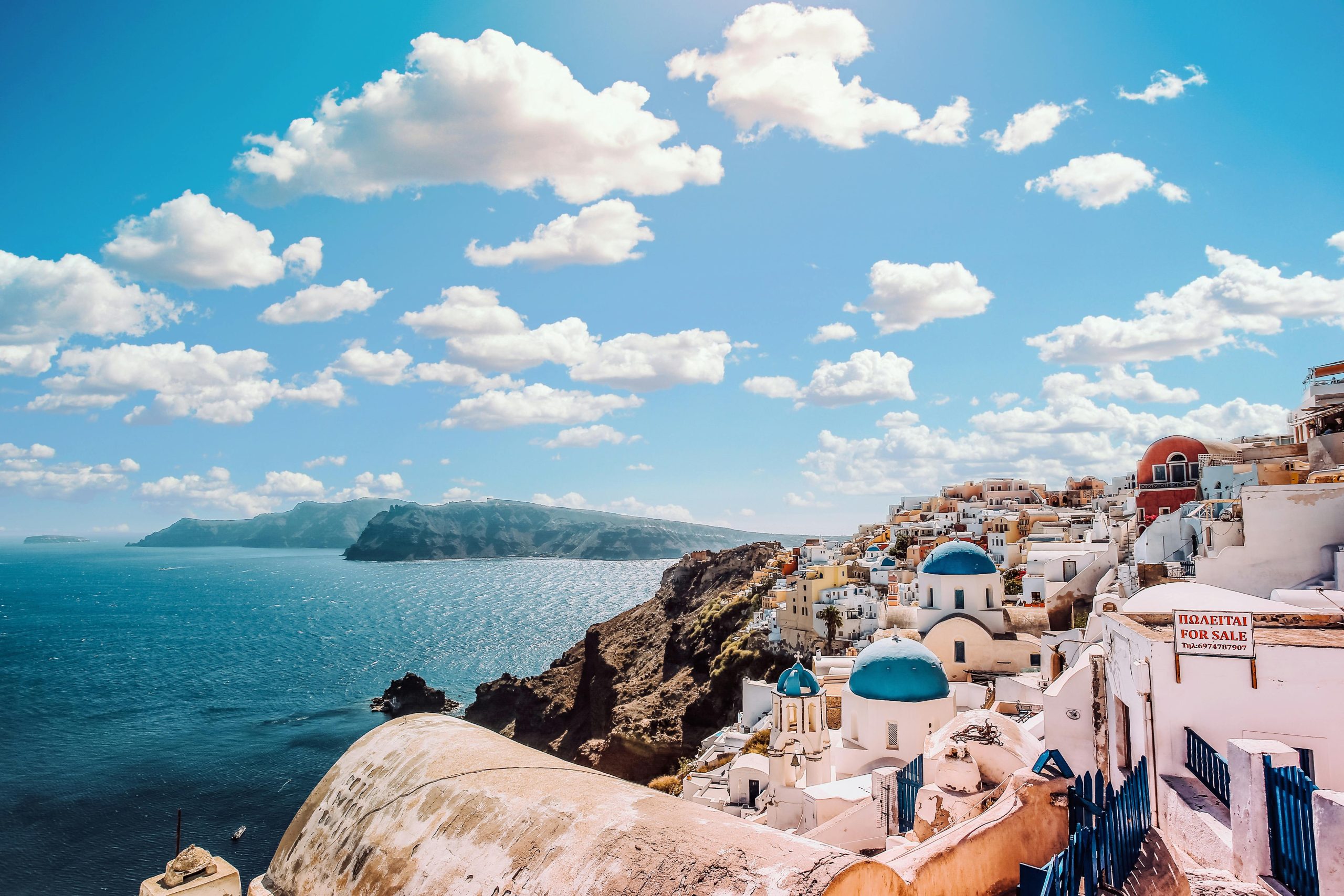 White-washed buildings with blue domed roofs overlook the crystal-clear Aegean Sea in Santorini, Greece, under a sky dotted with fluffy clouds