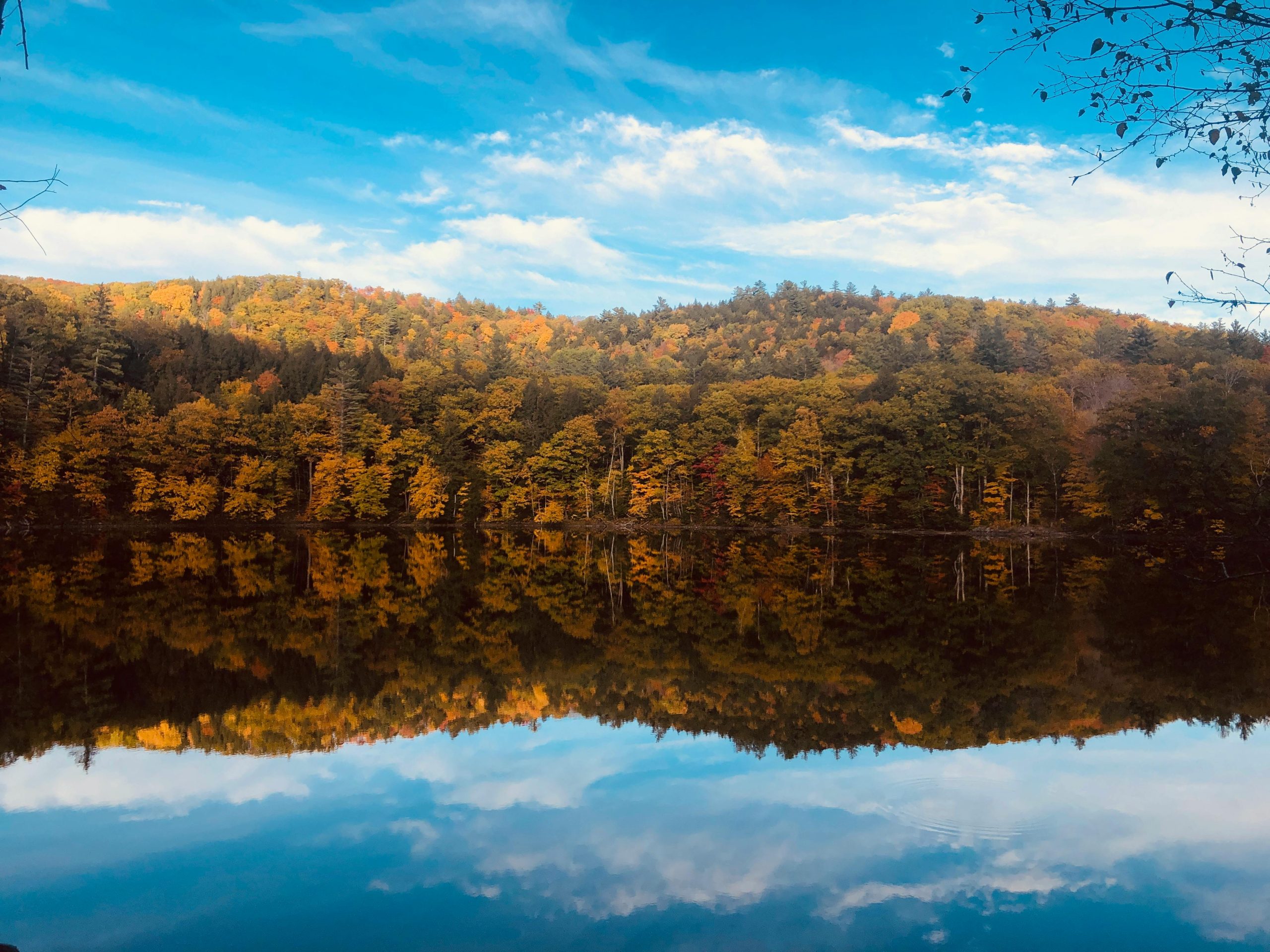 Calm, reflective lake surrounded by a dense forest of trees in full autumn colors, with the vibrant foliage mirrored perfectly on the water's surface under a blue, cloud-streaked sky.