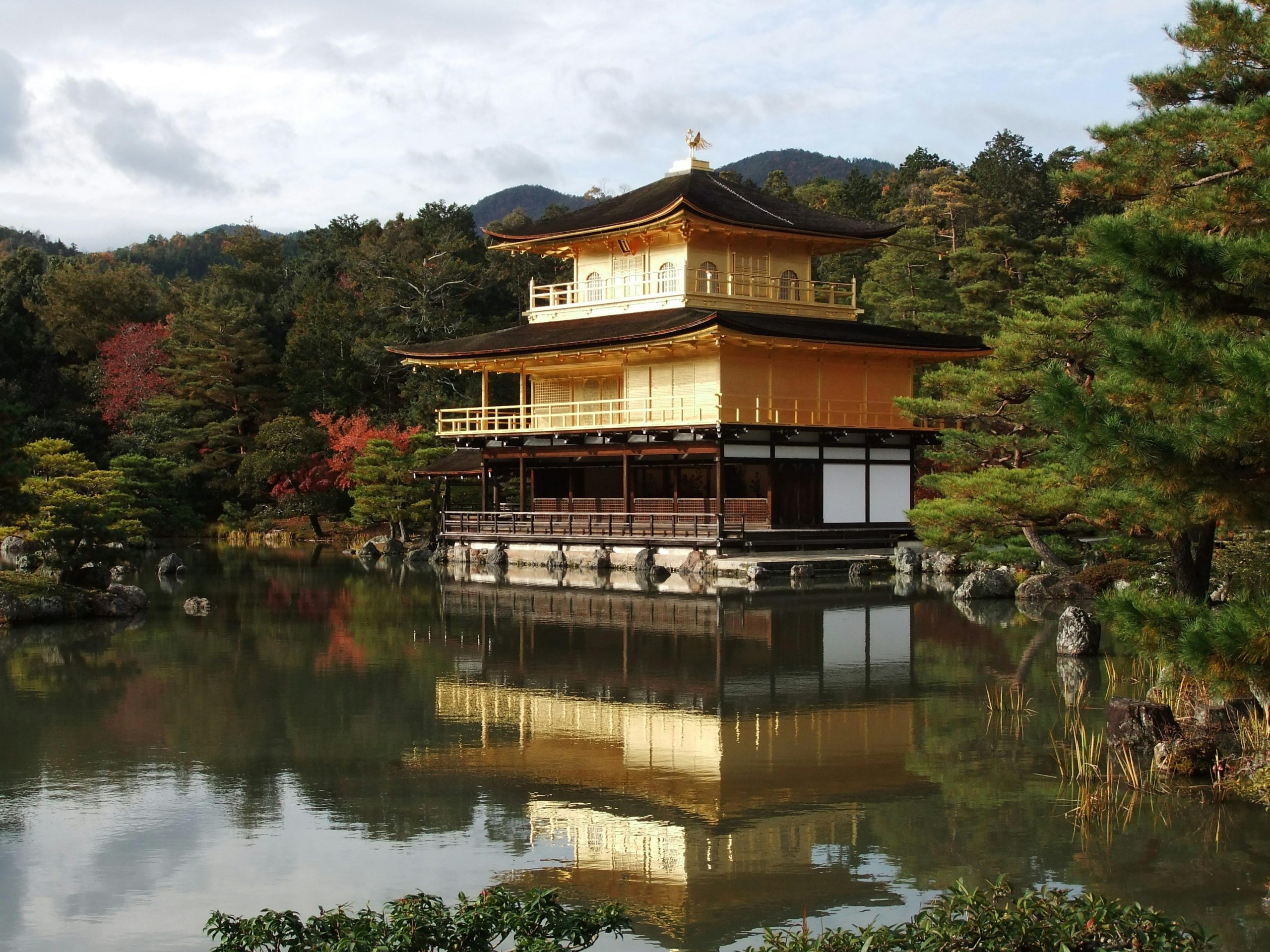 Golden Pavilion (Kinkaku-ji) reflected on the calm waters of a surrounding pond, set against a backdrop of lush trees and distant mountains. The golden structure shines in the sunlight, creating a serene and picturesque scene.