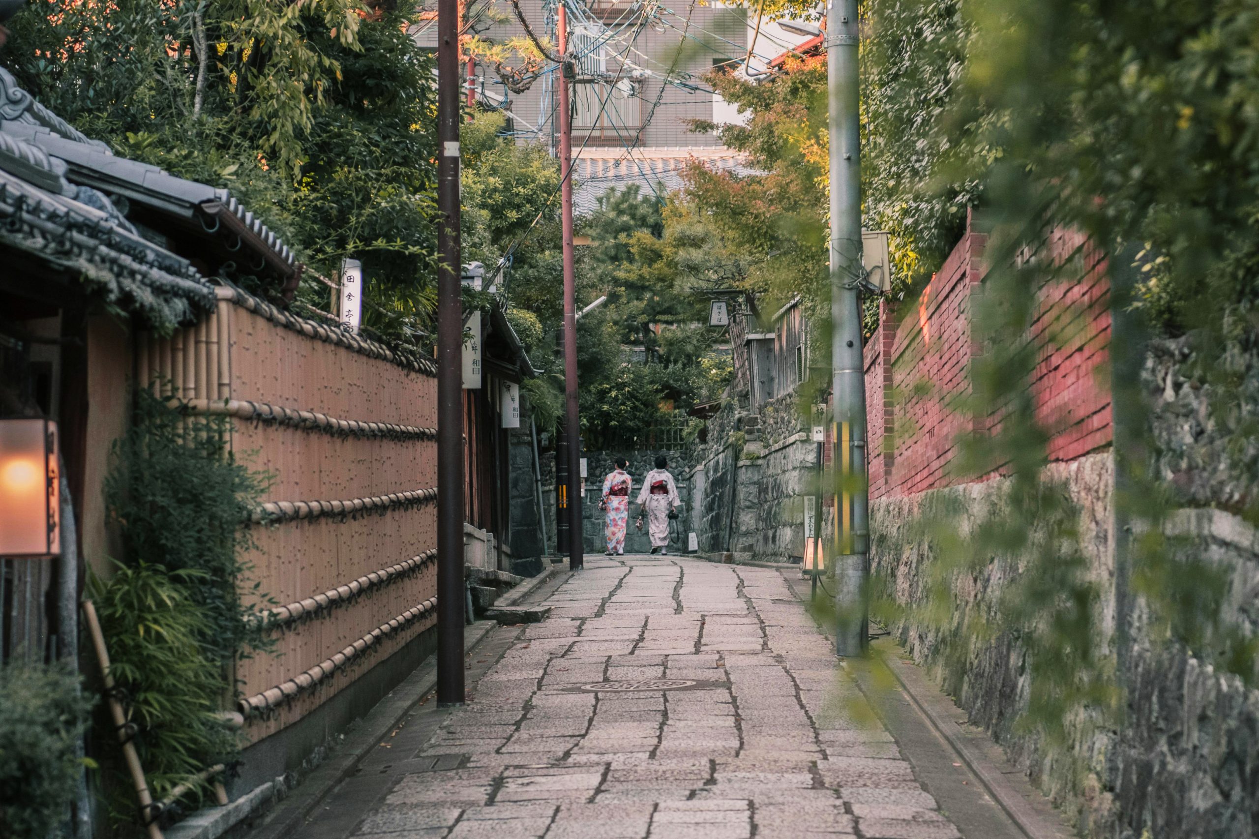 Two people dressed in traditional kimonos walk down a quiet, narrow alleyway in Kyoto, Japan, surrounded by serene, historic architecture and lush greenery.