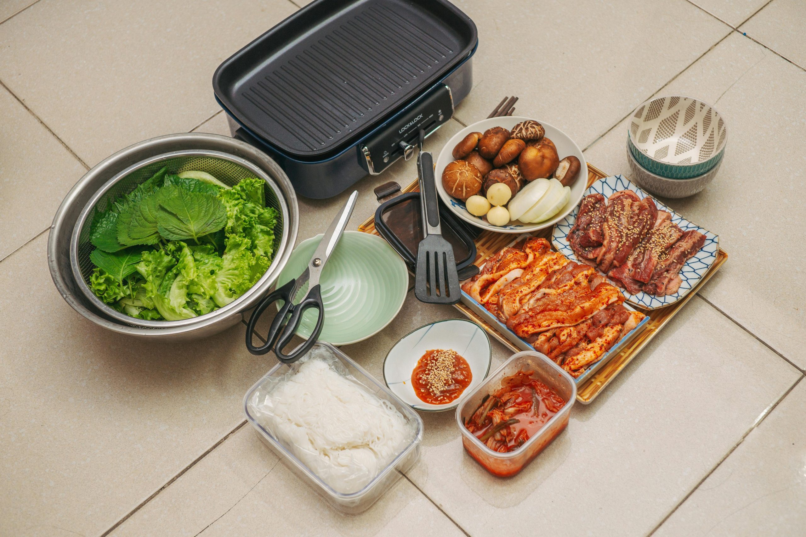 A variety of ingredients prepared for a Korean barbecue meal. The setup includes marinated meats, such as beef and chicken, ready to be grilled, alongside mushrooms, lettuce, and perilla leaves for wrapping. There are bowls of rice noodles, kimchi, and dipping sauce. A grill pan and utensils like scissors and spatulas are also visible, creating a cozy and delicious meal prep scene.