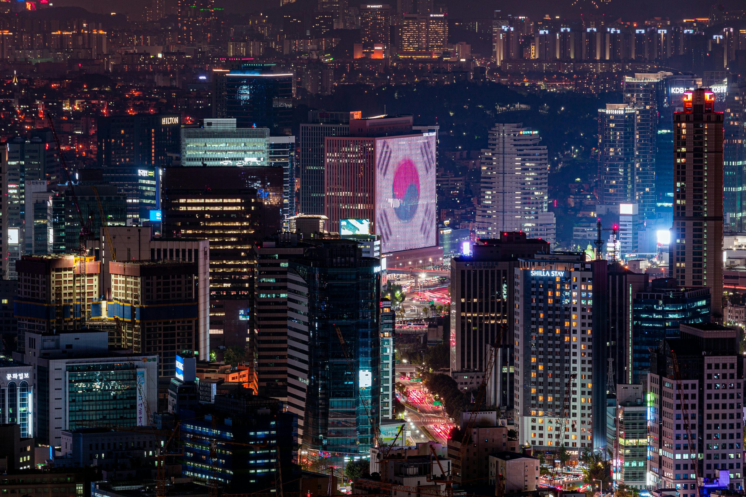 A vibrant cityscape of Seoul, South Korea, at night, featuring tall buildings and illuminated skyscrapers. In the foreground, a large digital billboard displays the South Korean flag, adding to the lively atmosphere. The city is bustling with lights, with streets lit up by car headlights and building lights. In the background, more skyscrapers and residential buildings are visible under the dark sky, giving a sense of the vast urban landscape.