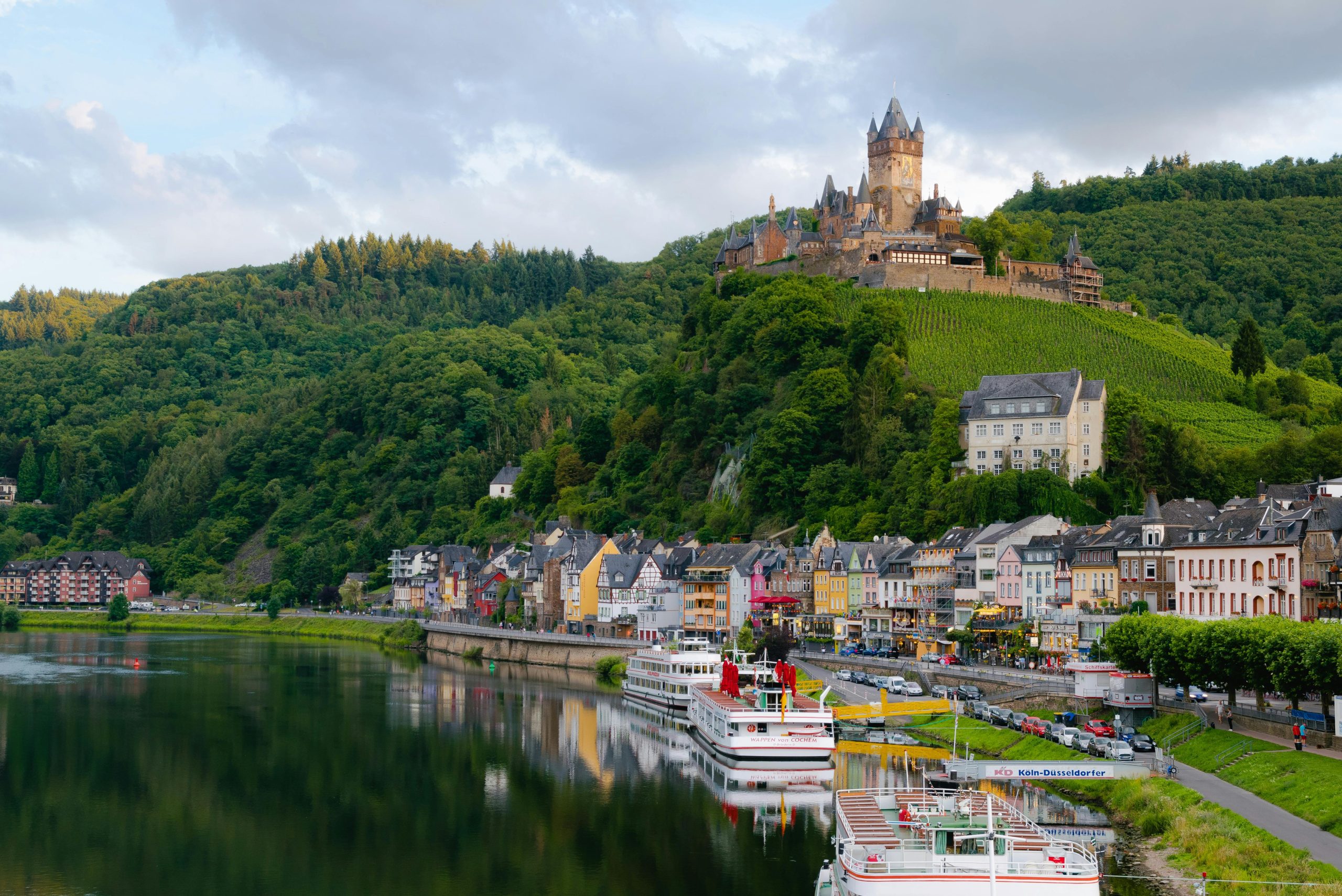 A picturesque riverside scene showcasing colorful buildings along a calm river, with a white boat docked in the foreground. On the hill above, a grand castle stands proudly, surrounded by lush greenery and vineyards. The sky is partly cloudy, casting a soft light over the charming town, while the calm waters reflect the vibrant buildings and the surrounding landscape, creating a serene and scenic view.