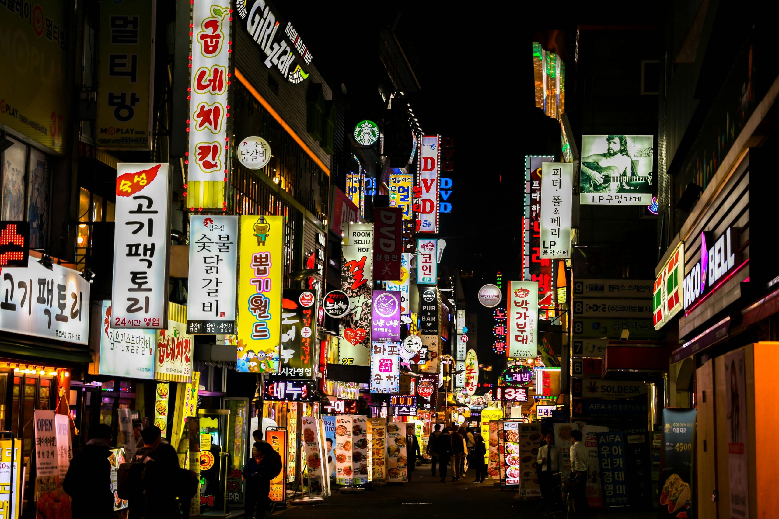 A vibrant, neon-lit street scene at night, filled with colorful signs in Korean, advertising various businesses like restaurants, bars, and shops. The bright lights from the signs create a lively and bustling atmosphere. Some familiar brands like Starbucks and Taco Bell are visible among the local establishments. A few people can be seen walking down the street, adding to the lively vibe of the area. The dense collection of signs and the warm glow of the lights showcase the energy of the nighttime city scene.