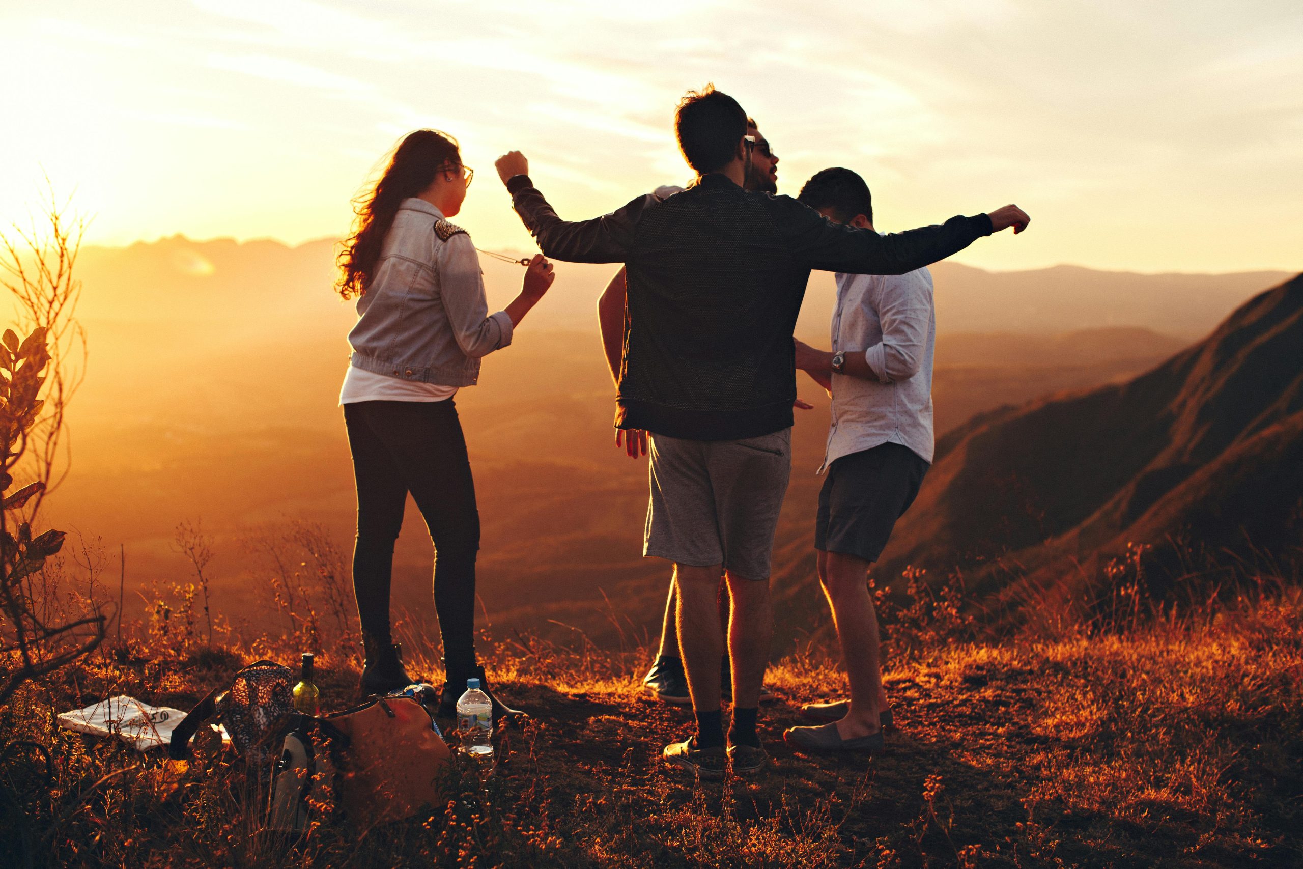 A group of four friends standing together on a mountaintop at sunset, enjoying the view. The scene captures a warm, golden glow as they embrace the moment, with one person holding a camera while others chat and laugh. They are casually dressed, surrounded by nature, with backpacks and bottles placed on the ground. The peaceful, scenic landscape in the background enhances the sense of adventure and camaraderie.