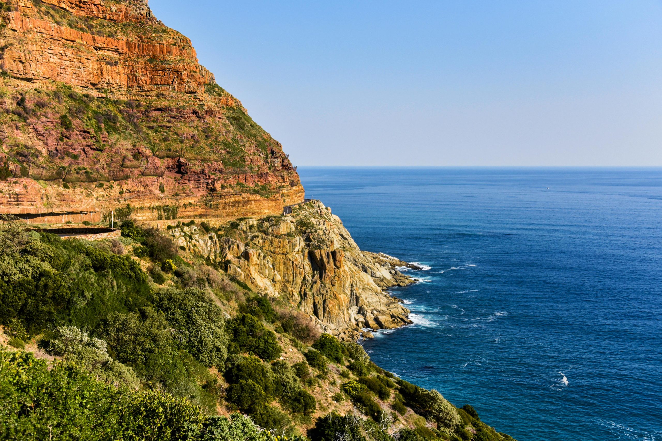 Scenic coastal view of a rugged cliff covered in lush greenery and colorful rock layers, overlooking the deep blue ocean under a clear, sunny sky.