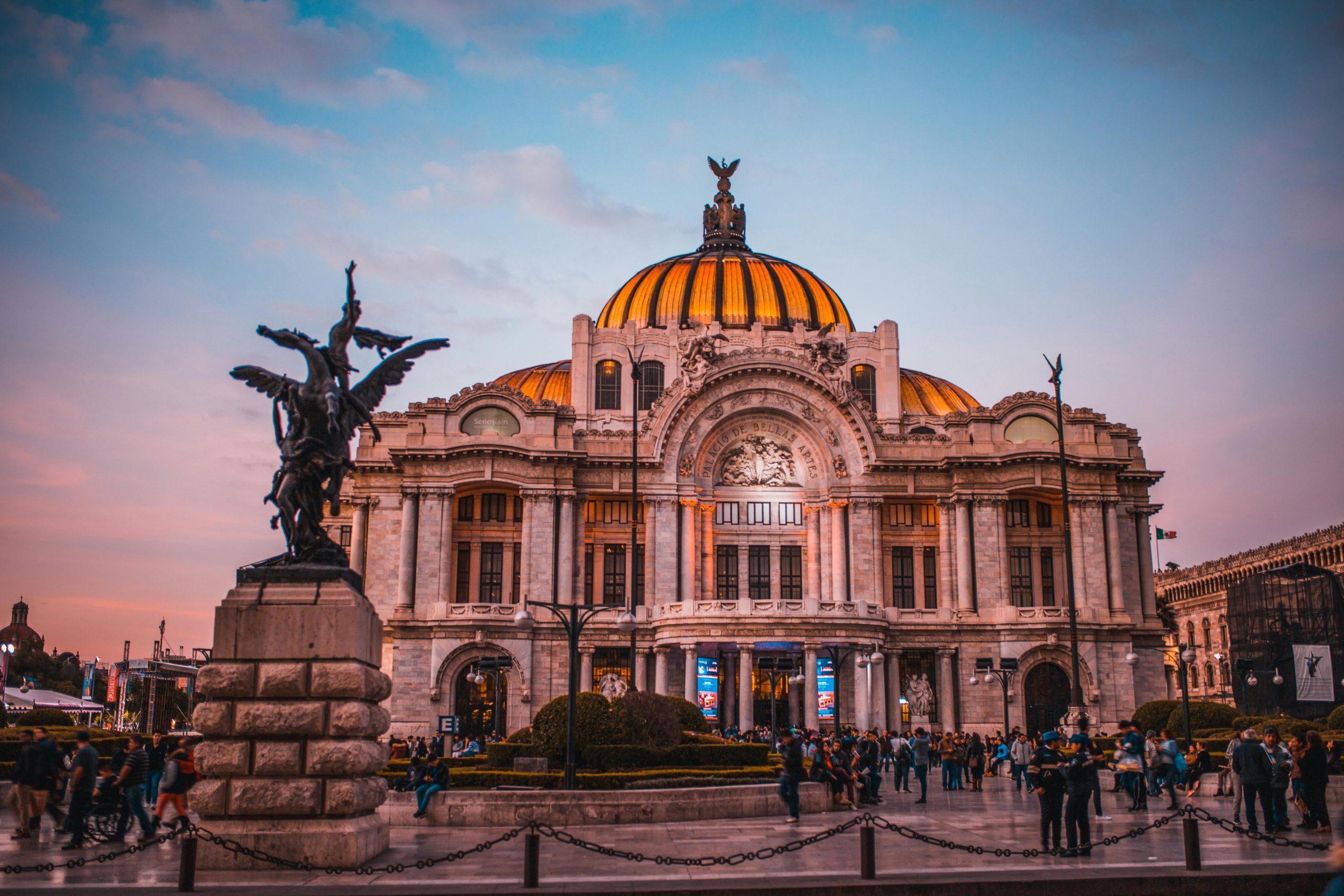 Stunning view of the Palacio de Bellas Artes in Mexico City during sunset, showcasing its iconic golden dome, intricate architectural details, and the bustling square with visitors and a winged statue in the foreground.