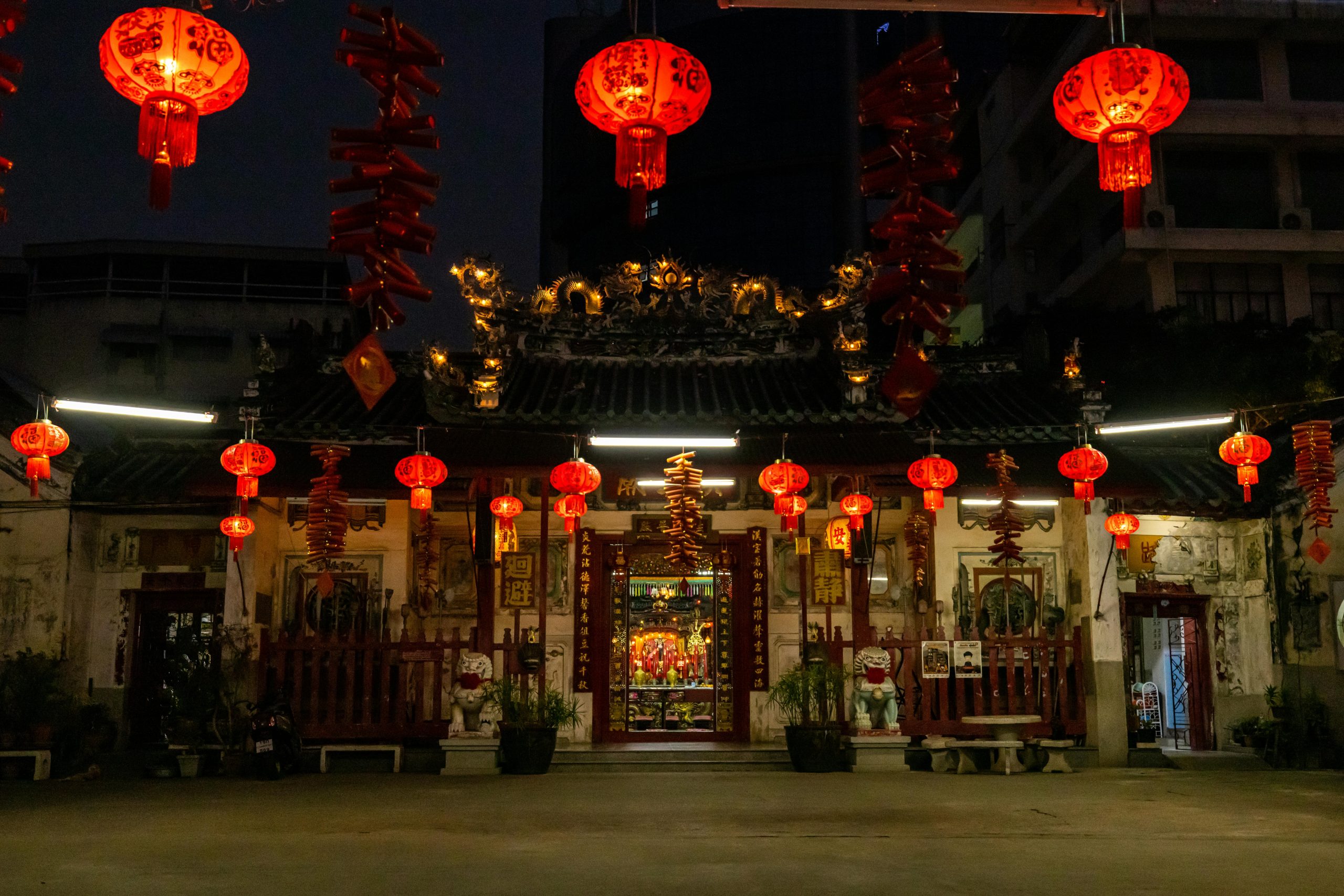A traditional Chinese temple beautifully illuminated at night with red lanterns hanging around its entrance. The temple's roof features intricate details, including dragon carvings, while the surrounding area is decorated with hanging red symbols and decorations. In the foreground, two guardian lion statues stand at the entrance, adding to the cultural richness of the scene. The warm light from the lanterns creates a peaceful yet vibrant atmosphere, contrasting with the urban backdrop of modern buildings.