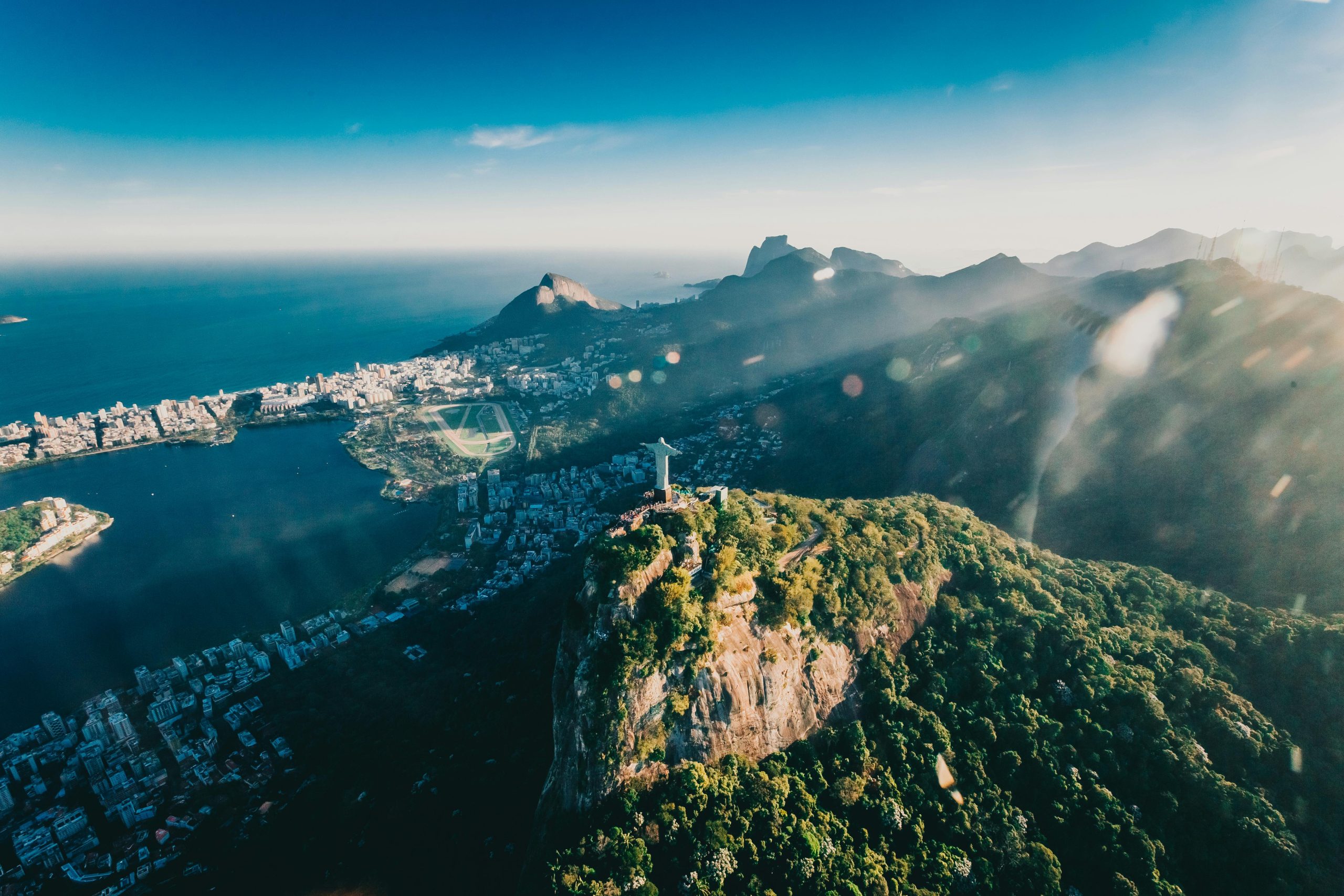 The image showcases a breathtaking aerial view of Rio de Janeiro, Brazil, with the iconic Christ the Redeemer statue atop the Corcovado Mountain. Below, the city stretches along the coastline, with a mix of urban buildings, lush greenery, and the serene blue ocean. The sunlight filters through the landscape, casting a warm glow over the mountainous terrain and cityscape. The image captures the beauty of both the natural and urban environments of Rio de Janeiro.