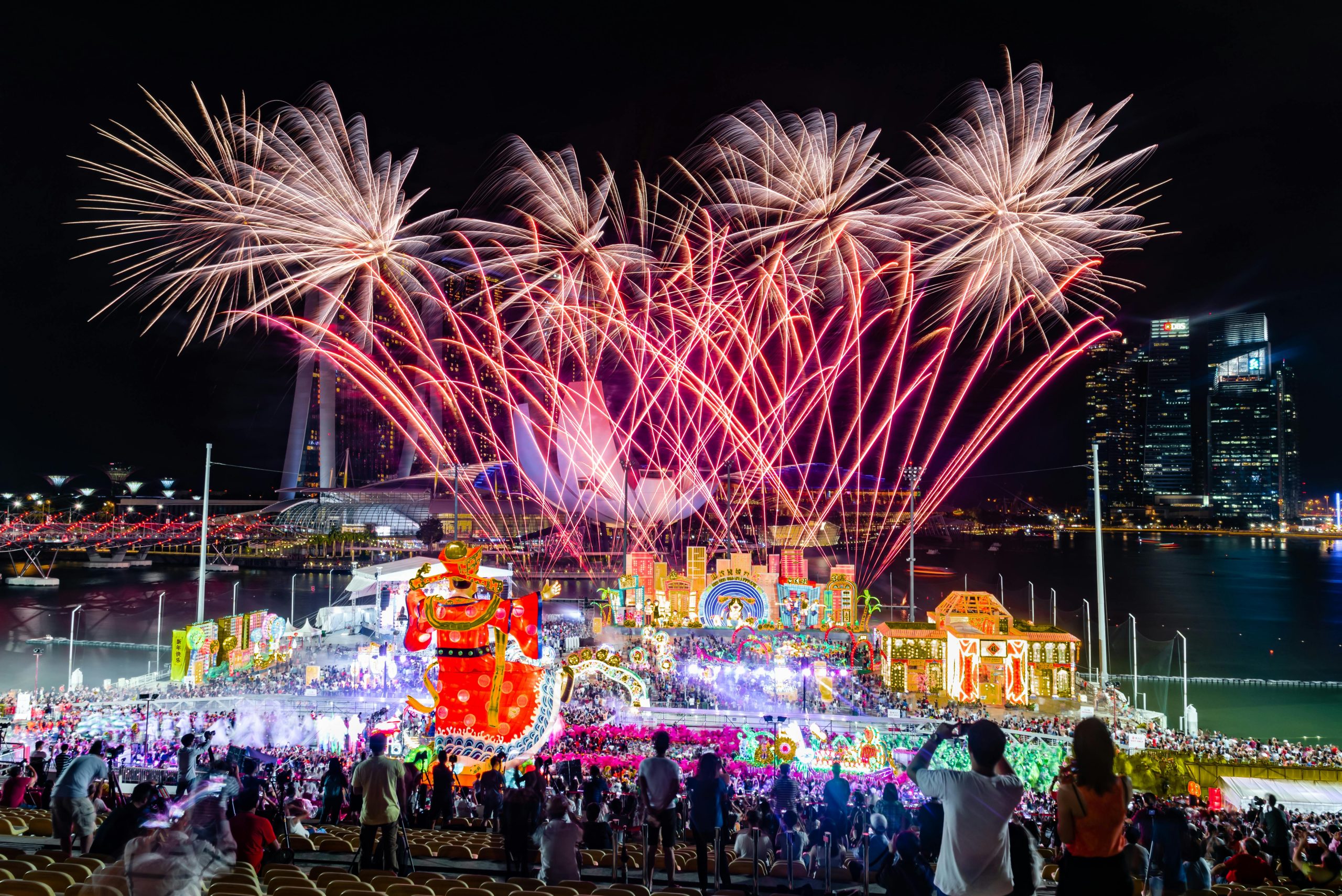 A vibrant night scene from a grand festival, with colorful fireworks exploding in the sky above a lively crowd. Below, a spectacular display of illuminated floats and large, brightly colored decorations creates a festive atmosphere, set against the backdrop of modern buildings by the water. The audience, seated in the foreground, eagerly watches the festivities unfold, capturing the magical moment on their phones. The celebration exudes energy, with lights and vibrant colors enhancing the festive spirit of the event.