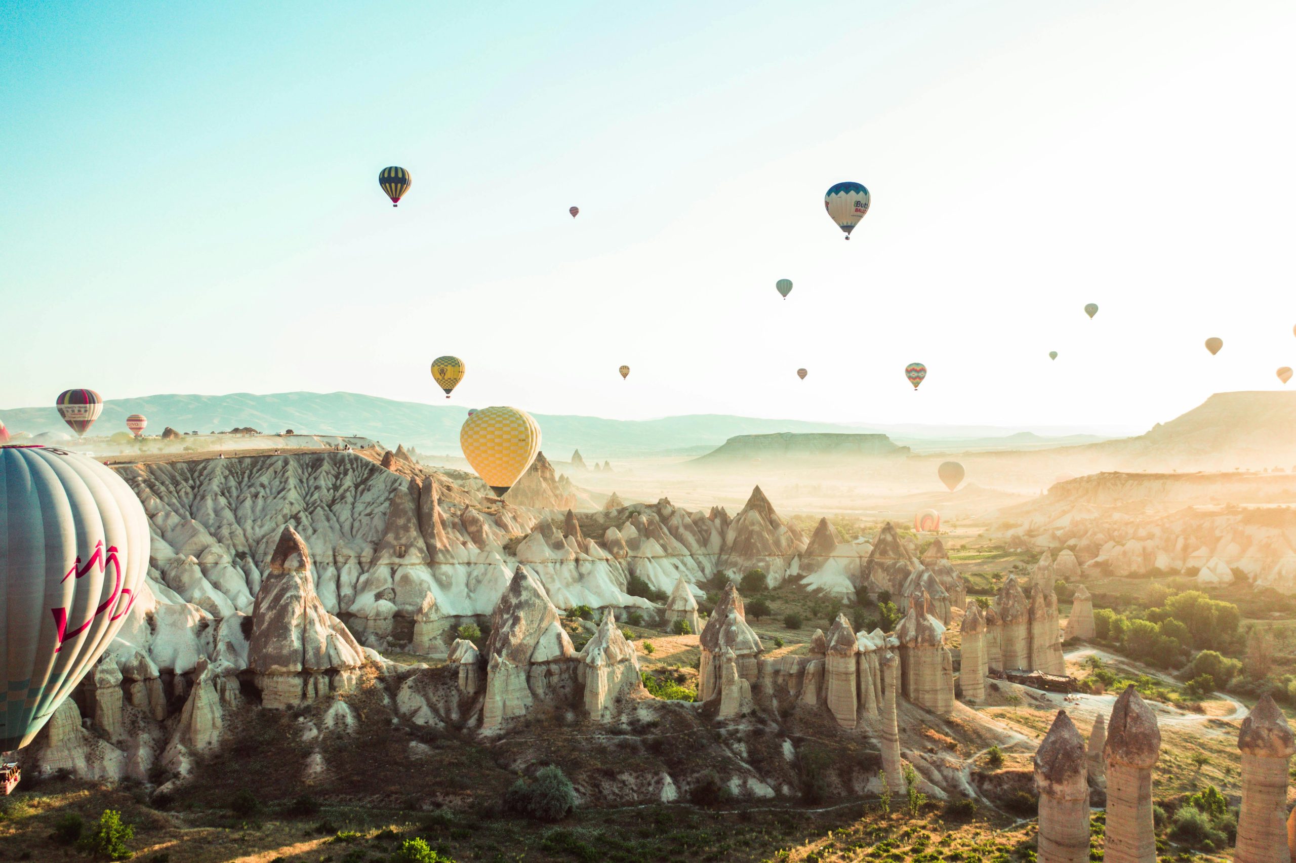 An aerial view of colorful hot air balloons floating over the unique, rocky landscape of Cappadocia, Turkey, at sunrise. The soft morning light illuminates the surreal formations of the region, creating a serene and magical atmosphere.