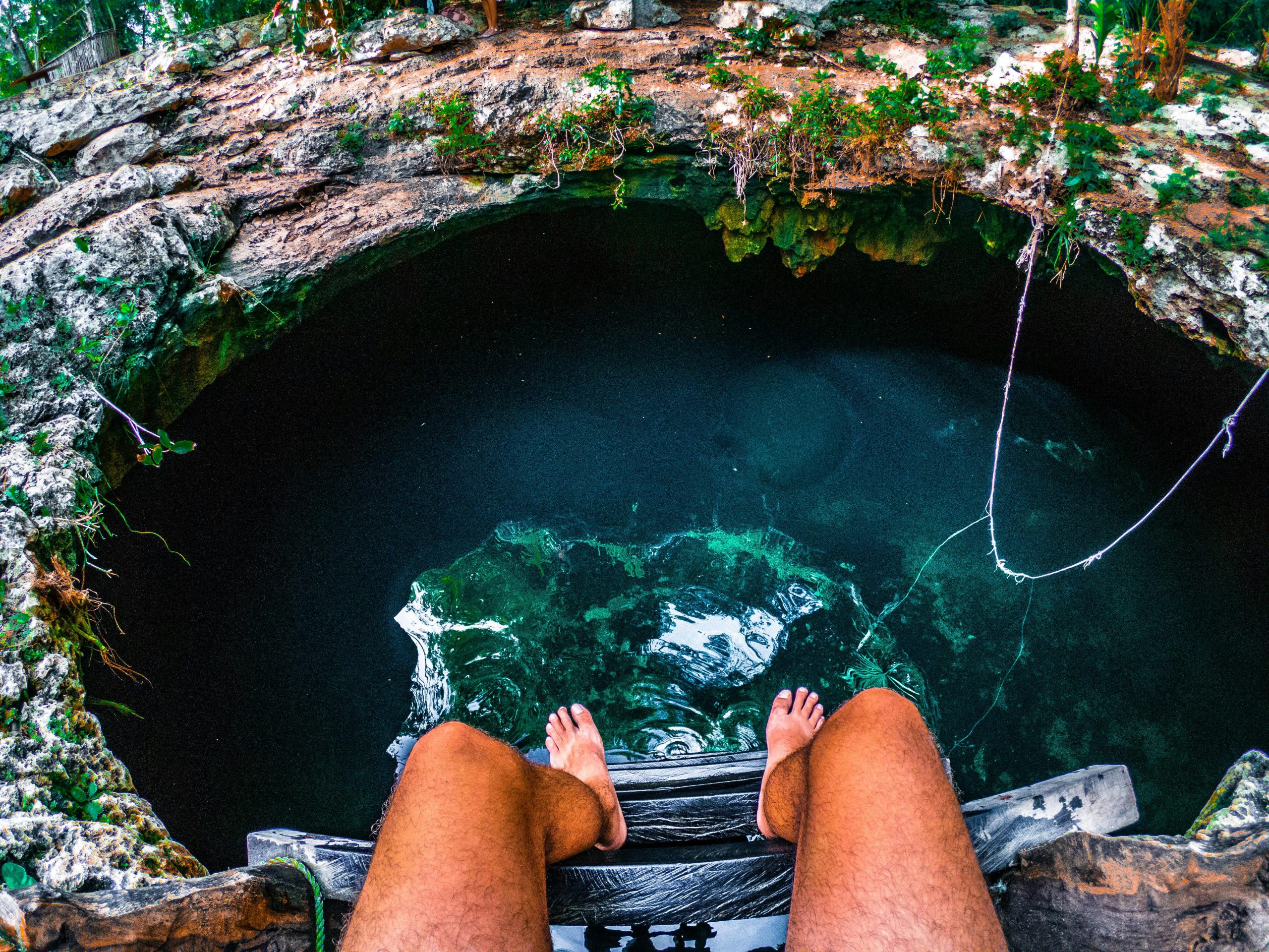 A person sitting on the edge of a wooden platform, with their feet dangling above a crystal-clear cenote, surrounded by lush greenery and rocky terrain. A rope hangs nearby, adding to the adventurous atmosphere of the natural swimming hole.