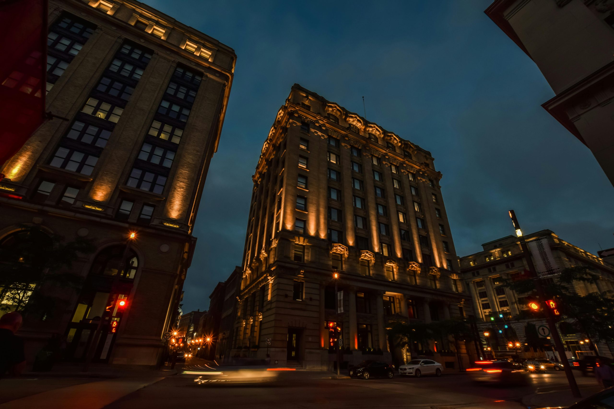 View of two tall, illuminated buildings at dusk, with their architectural details highlighted by warm lights. The streets below are lit up with streetlights, and a car drives past, leaving streaks of light as the city comes to life in the evening.