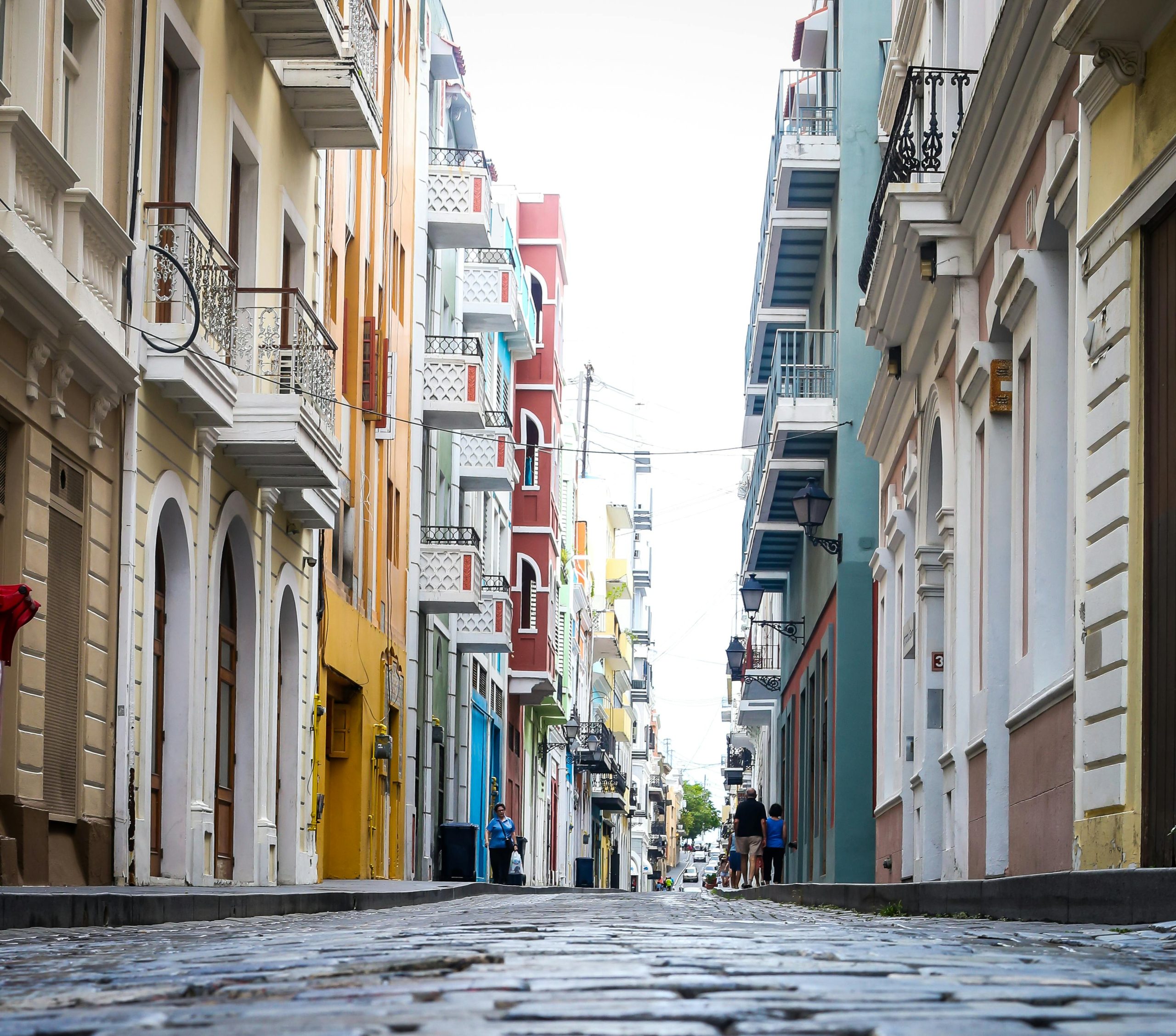 A narrow, cobblestone street in a colorful, historic neighborhood, with vibrant buildings in shades of yellow, red, pink, and blue. The street is lined with balconies, and a few people walk casually down the road, capturing the lively, charming atmosphere of the area.