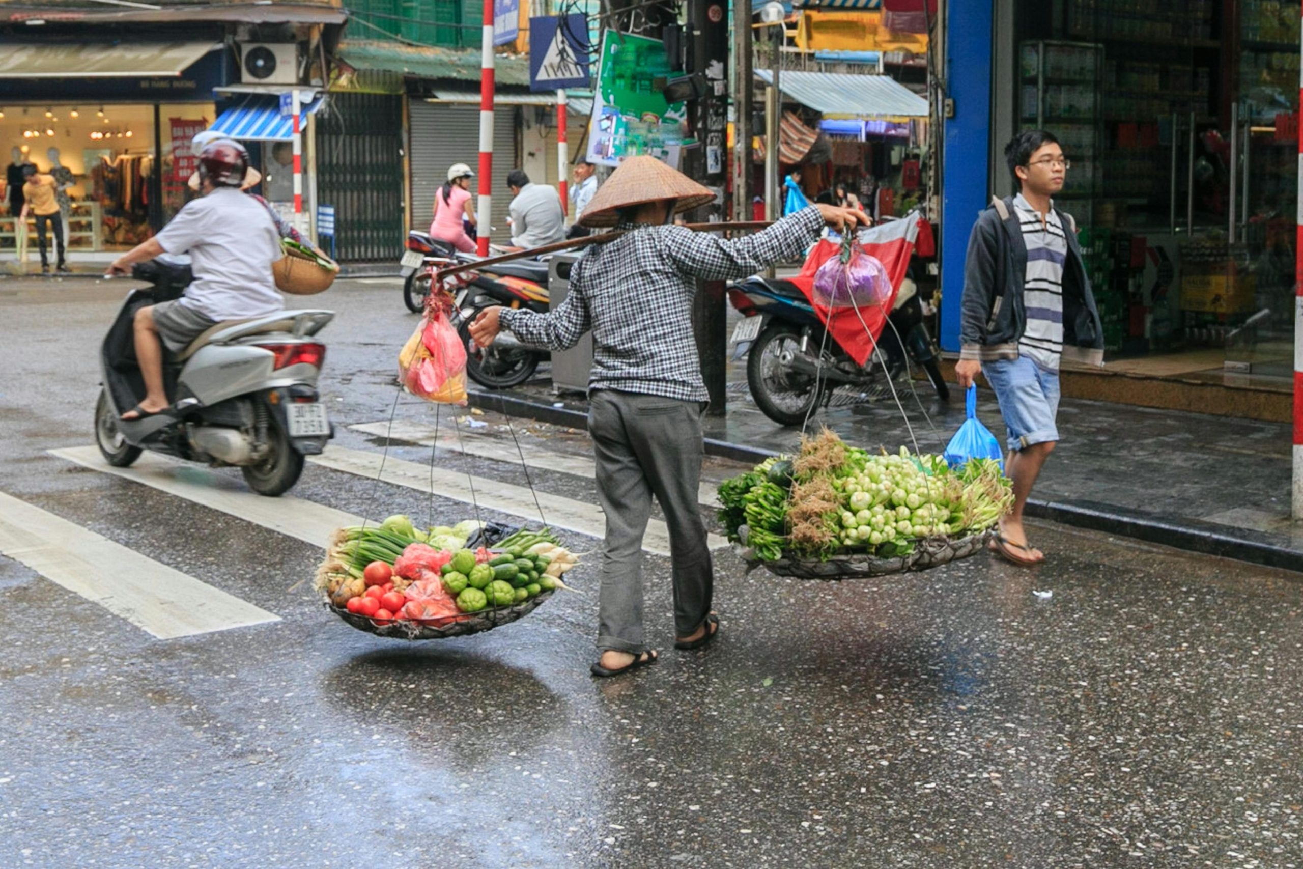 A street vendor in Vietnam carrying two baskets of fresh produce across a busy road, with colorful bags hanging from each side. Motorbikes pass by as pedestrians walk in the rain, capturing a lively scene in the heart of the city.
