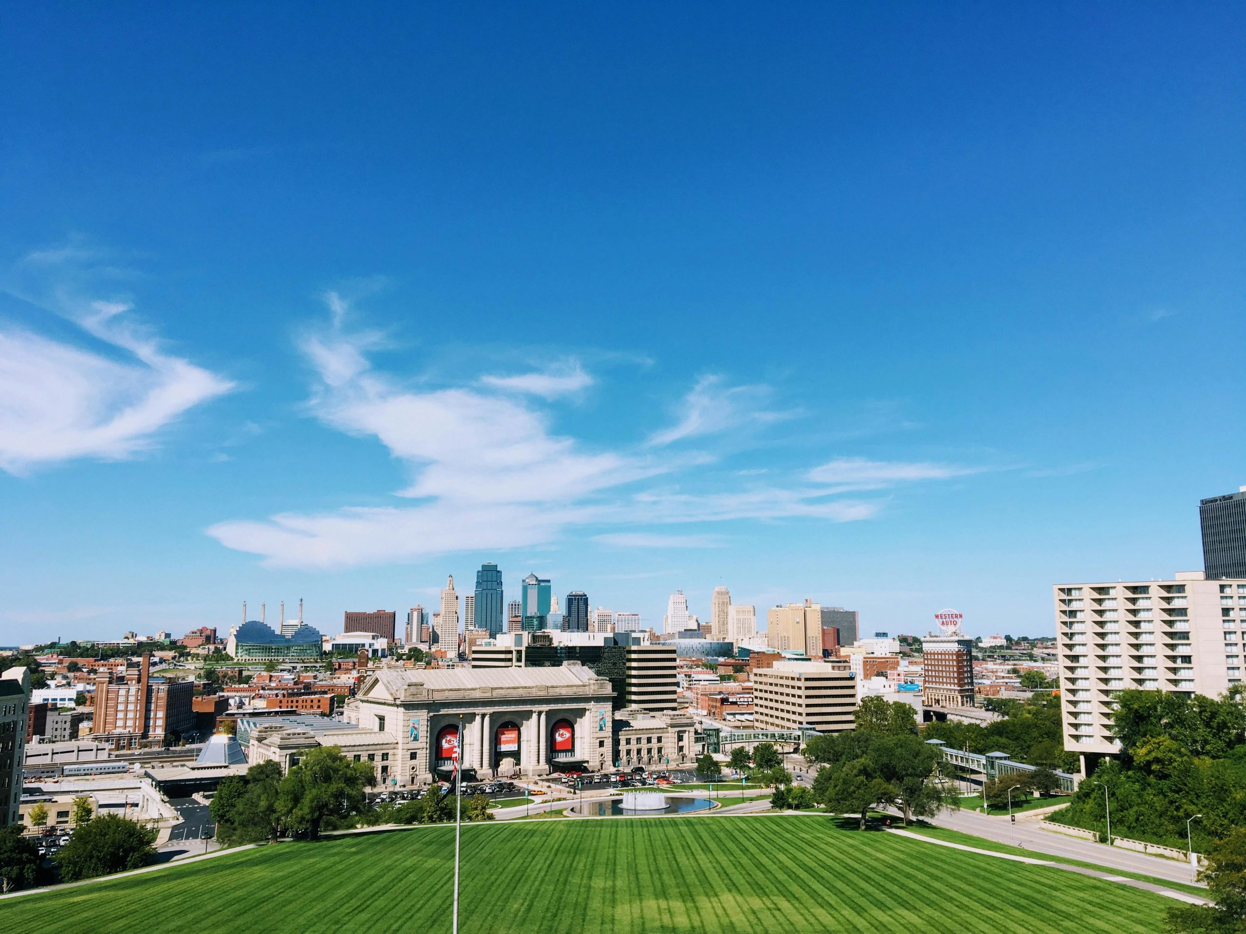 A panoramic view of the skyline of Kansas City with a clear blue sky. The image features tall buildings, including the distinctive Kansas City Power and Light Building and the historic Union Station, with lush green grass and tree-lined streets in the foreground.