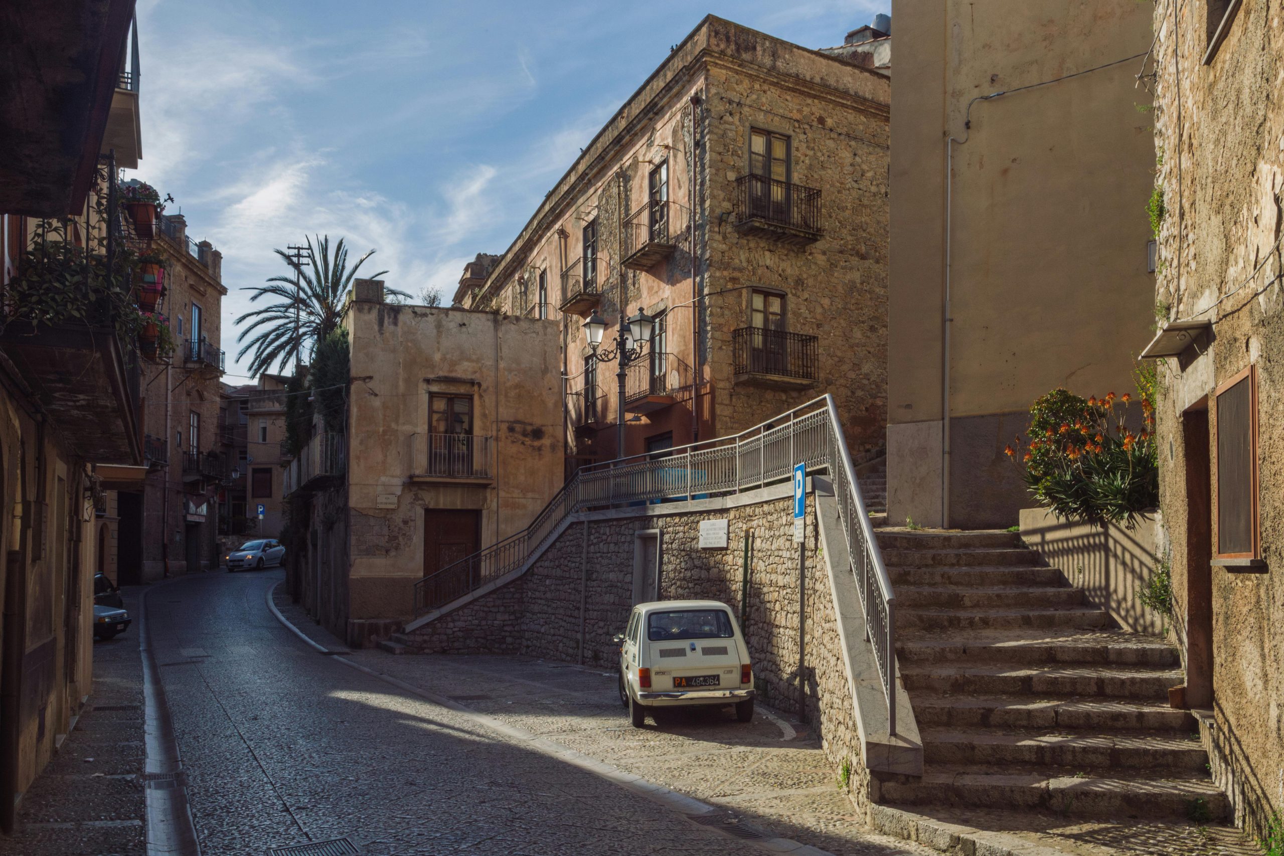 A quiet cobblestone street in a charming European village, lined with rustic stone buildings, a staircase with a metal railing, and a vintage white car parked near the curb under a clear blue sky