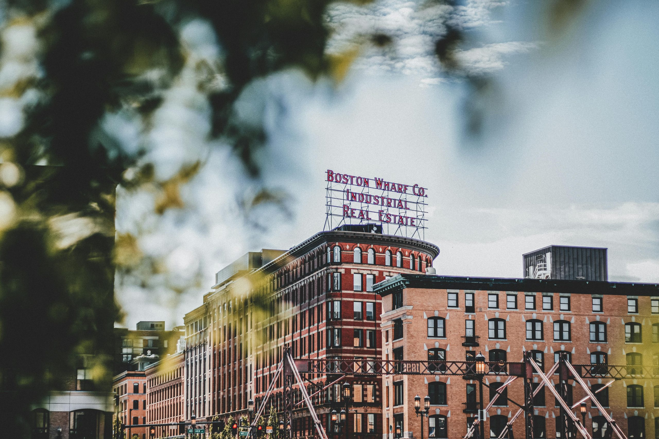 View of a historic building in Boston with the 'Boston Wharf Co. Industrial Real Estate' sign at the top. The photo captures the architectural charm of the brick building, partially framed by foliage in the foreground.