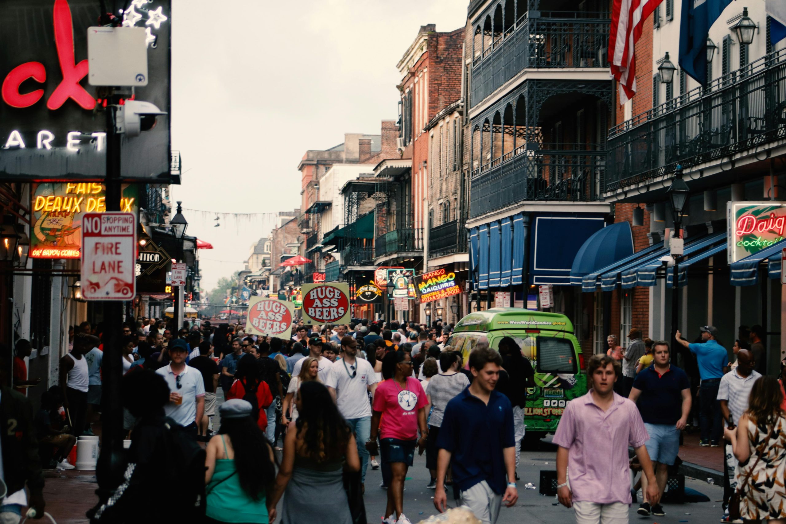 Busy street scene in New Orleans with crowds of people walking along a vibrant, bustling neighborhood. Neon signs for bars and restaurants illuminate the street, with vendors holding large signs while colorful architecture and balconies line the buildings.