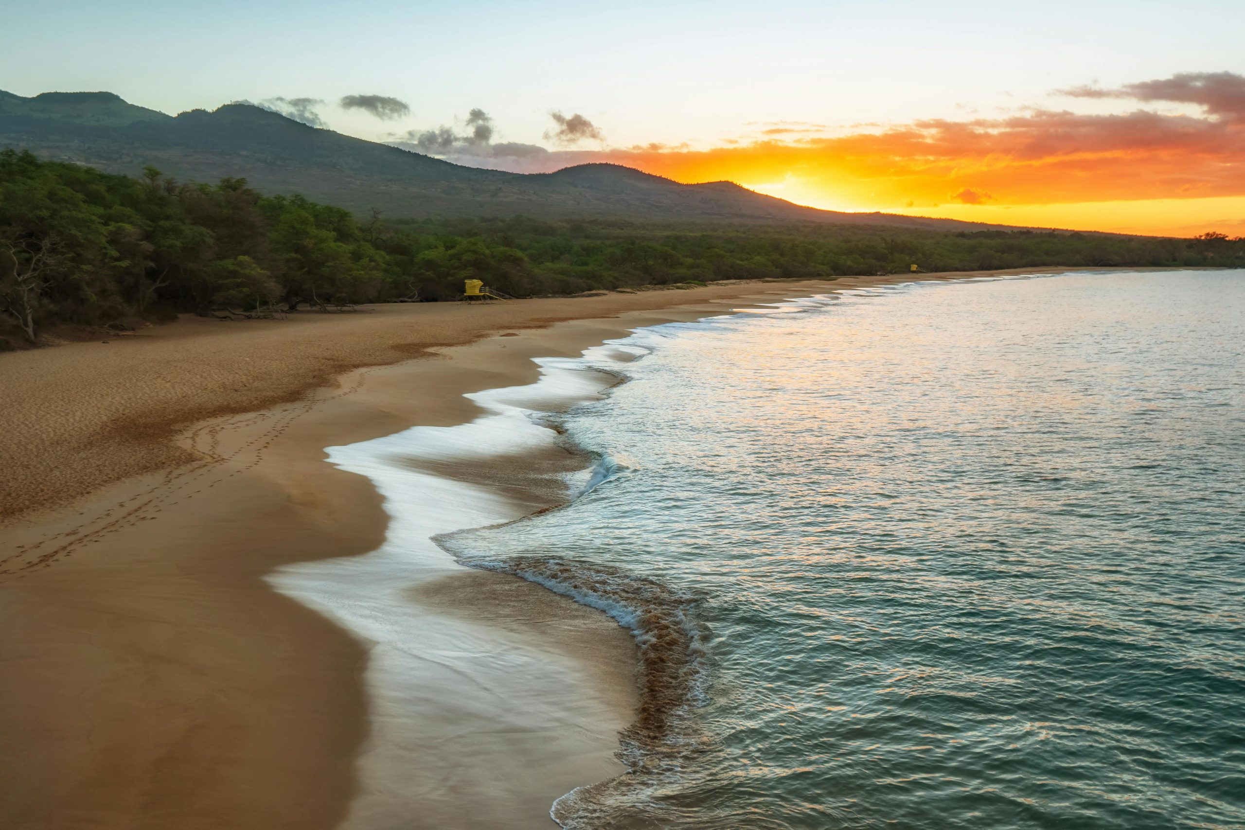 A serene beach at sunset with gentle waves washing onto the sandy shore, lush green trees lining the beach, and mountains in the distance under a colorful sky