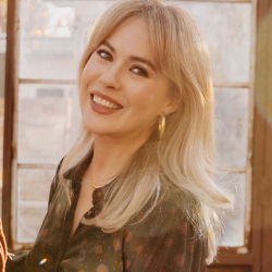 Portrait of a smiling blonde woman with shoulder-length hair, wearing a dark top, conveying a warm and approachable personality.