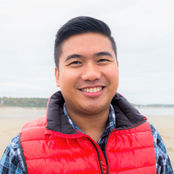 Portrait of a smiling man wearing a red vest outdoors near a body of water, conveying a friendly and approachable presence.