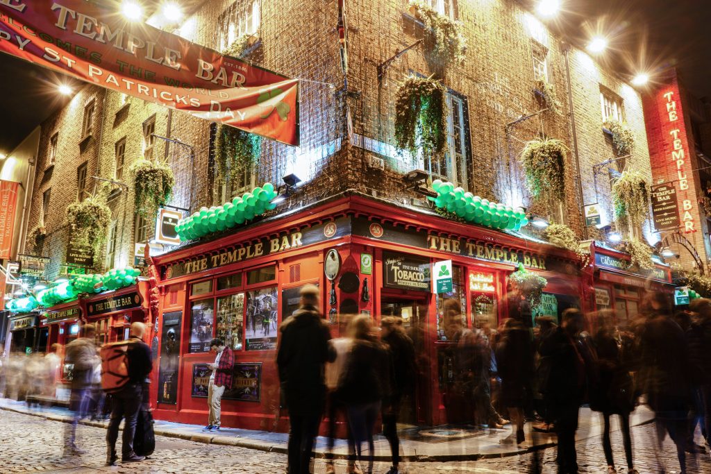 A vibrant, bustling scene outside The Temple Bar in Dublin, Ireland, adorned with St. Patrick’s Day decorations. Green balloons and lively crowds fill the area as people gather to celebrate.