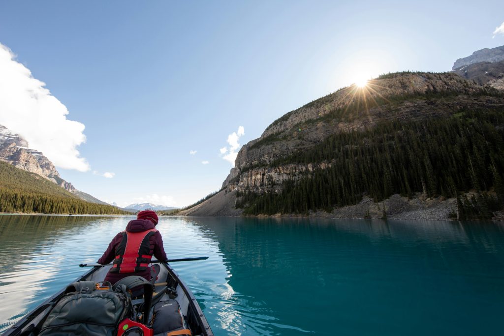 A solo kayaker paddles across a serene, turquoise lake surrounded by towering pine-covered mountains under a bright, clear sky with the sun peeking over the ridge.
