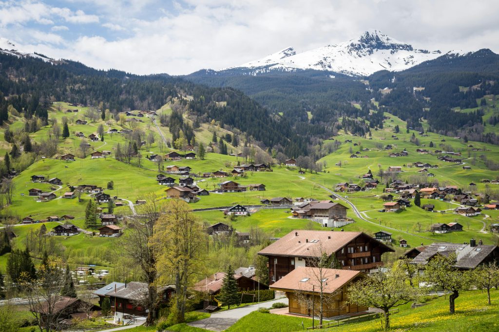 A picturesque view of a Swiss village nestled in the green hills with traditional wooden chalets scattered across the landscape. Snow-capped mountains rise majestically in the background under a cloudy sky.