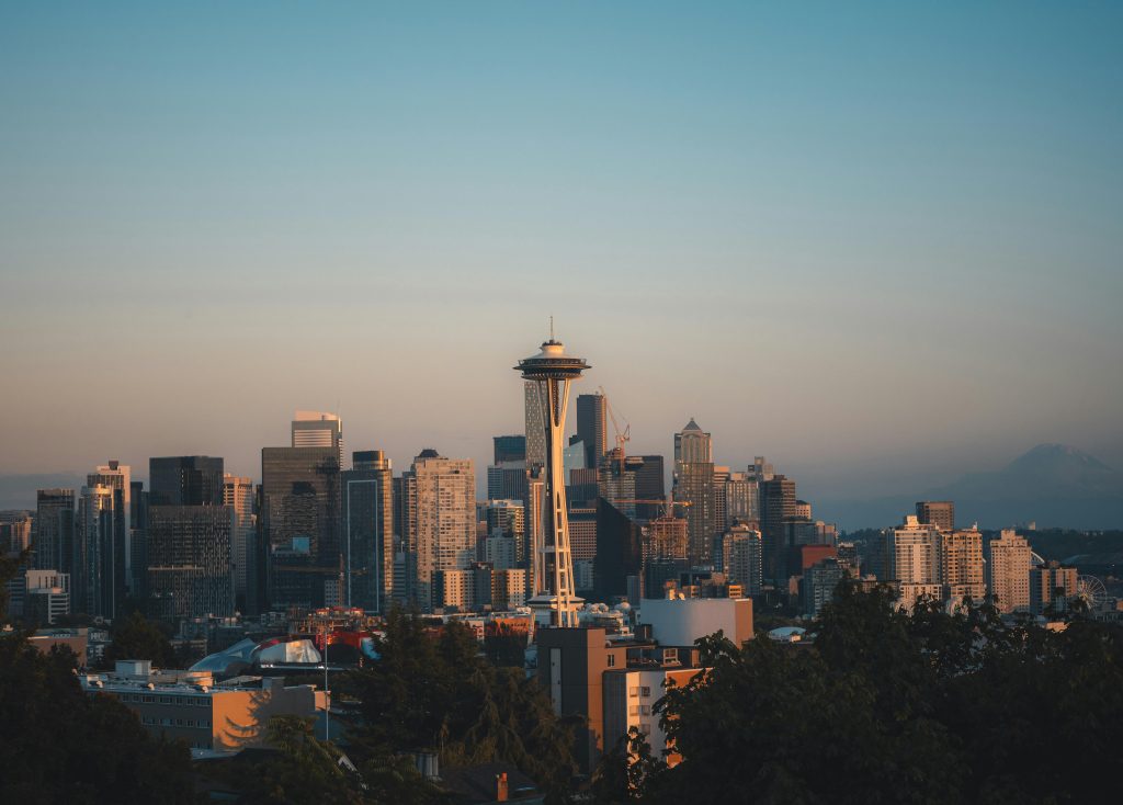 The Seattle skyline at dusk featuring the iconic Space Needle prominently in the foreground, with modern skyscrapers and Mount Rainier faintly visible in the distance.