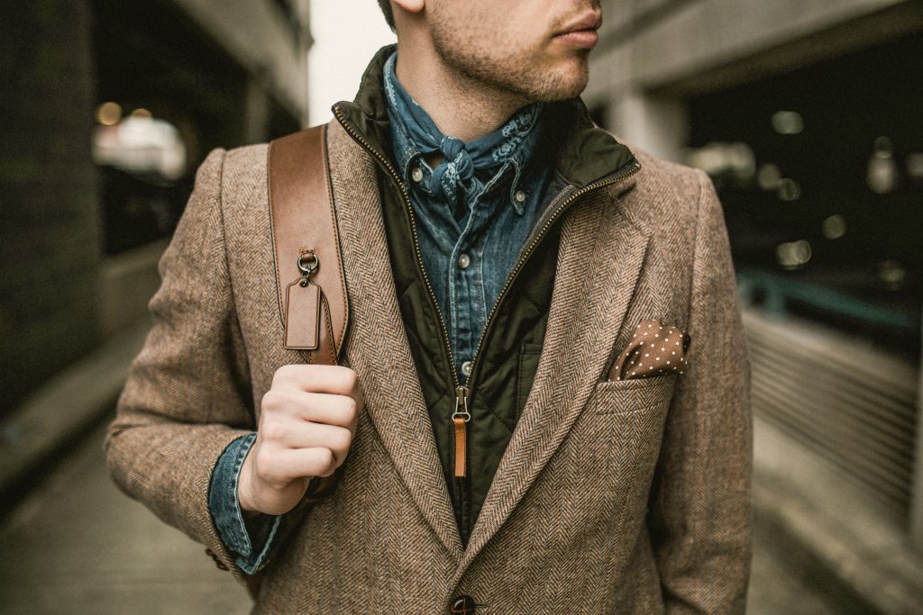 A stylish man wearing layered clothing, including a brown tweed blazer, a denim shirt, and a scarf, carrying a brown leather strap bag over his shoulder in an urban setting.