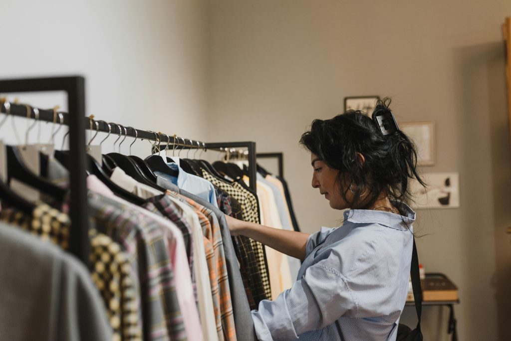 A young woman flips through shirts on a neatly organized clothing rack inside a cozy boutique. Soft, neutral lighting and varied patterns on the garments create a calm, curated shopping vibe.
