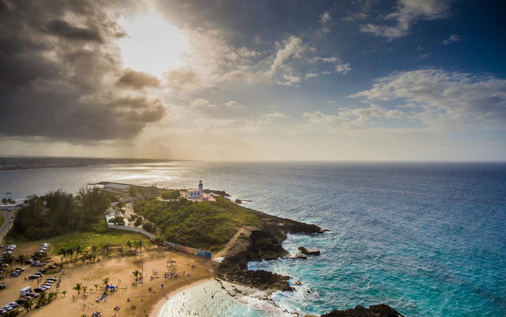 A scenic aerial view of a beach and rocky coastline with turquoise waters, a small lighthouse on a green hilltop, and people enjoying the sandy shore under a partly cloudy sky at sunset.