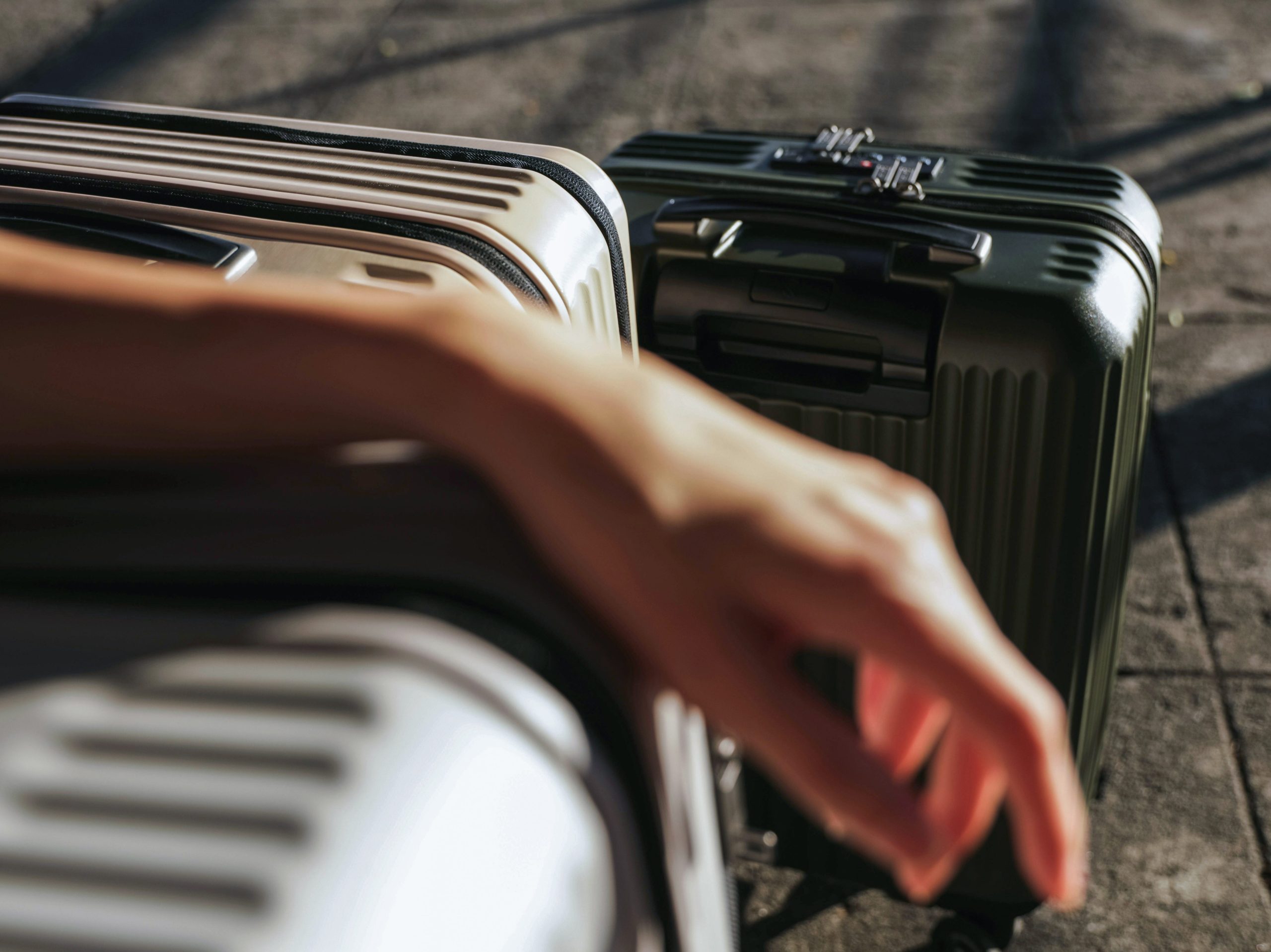 A close-up of a person’s hand resting on a beige hard-shell suitcase, with another dark green suitcase beside it on a sunlit sidewalk.