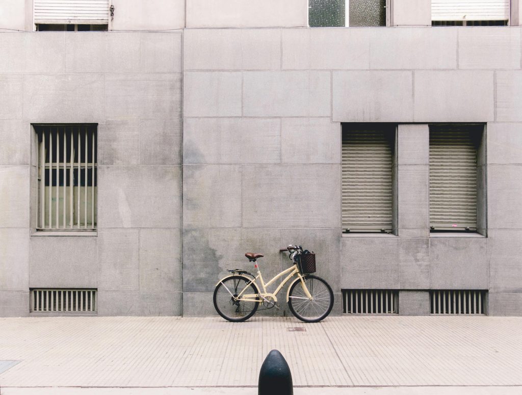 A cream-colored bicycle with a basket rests against a minimalist gray concrete building façade. The stark wall and shuttered windows emphasize the bike’s vintage charm and solitary presence.