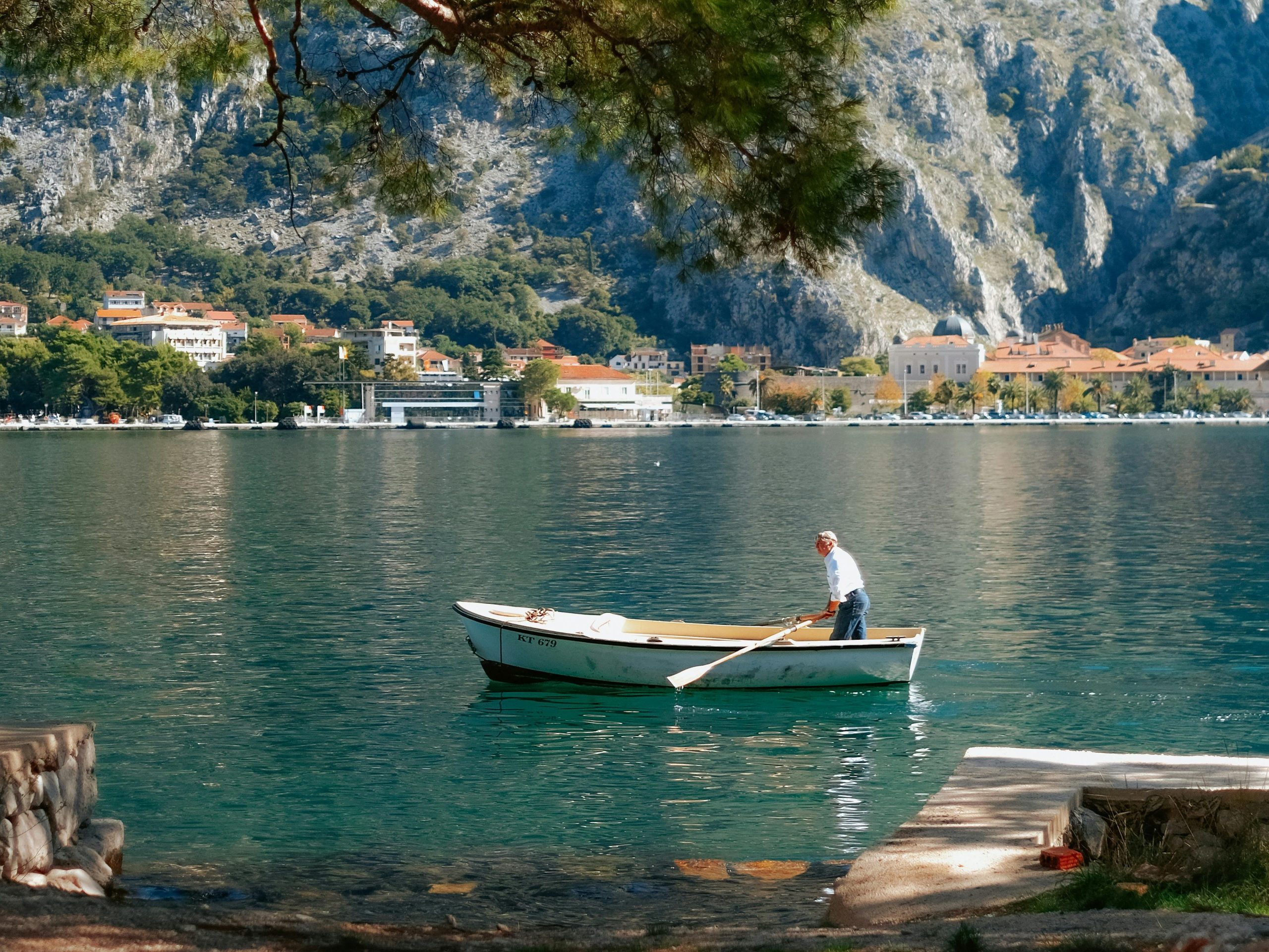 Man rowing a small boat on the Bay of Kotor, Montenegro, with a coastal village and rocky Dinaric Alps in the background — a hidden gem for summer travel in Europe.