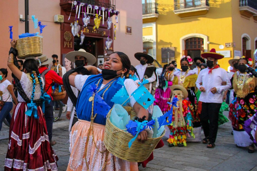 A vibrant street parade featuring people in traditional Mexican clothing, with women carrying decorated baskets and wearing colorful dresses, while others in the background wear sombreros and festive attire during a cultural celebration.