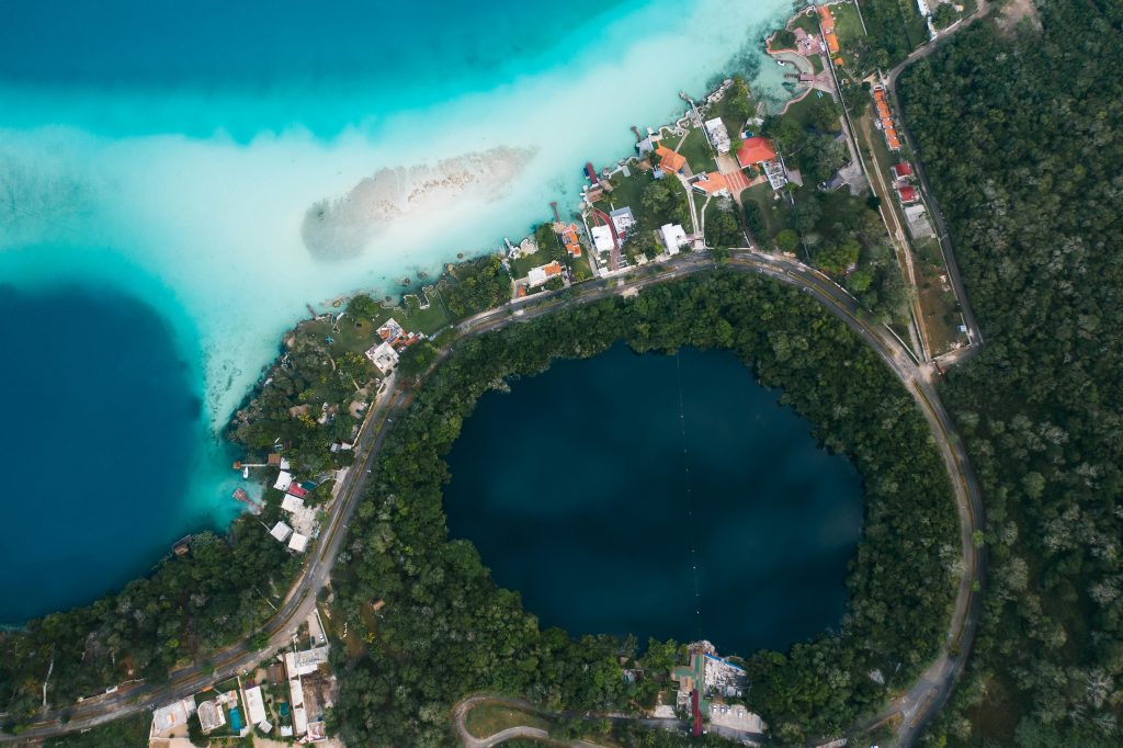 An aerial view of a vibrant blue and turquoise lagoon next to a dark circular cenote, surrounded by a lush forest and a small coastal town with houses and roads curving around the water.