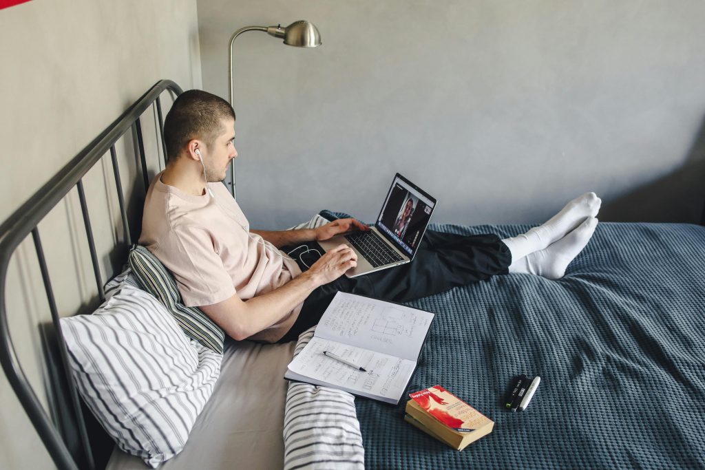 A college-age man reclines on his bed, earbuds in, focused on a video call on his laptop. Open notebooks and pens scattered around him show he’s multitasking between study and conversation.