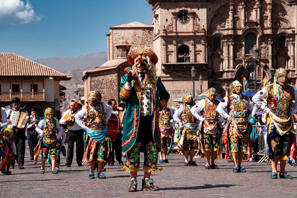A vibrant street parade in Cusco, Peru, featuring performers in traditional Andean costumes and colorful masks, dancing in front of historic colonial architecture with musicians playing instruments in the background.
