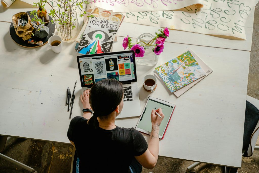 Viewed from above, a designer sketches lettering on a tablet at a large white table filled with art supplies, coffee, and vibrant magazines. The creative clutter and blooming flowers capture an inspiring workspace in full flow.